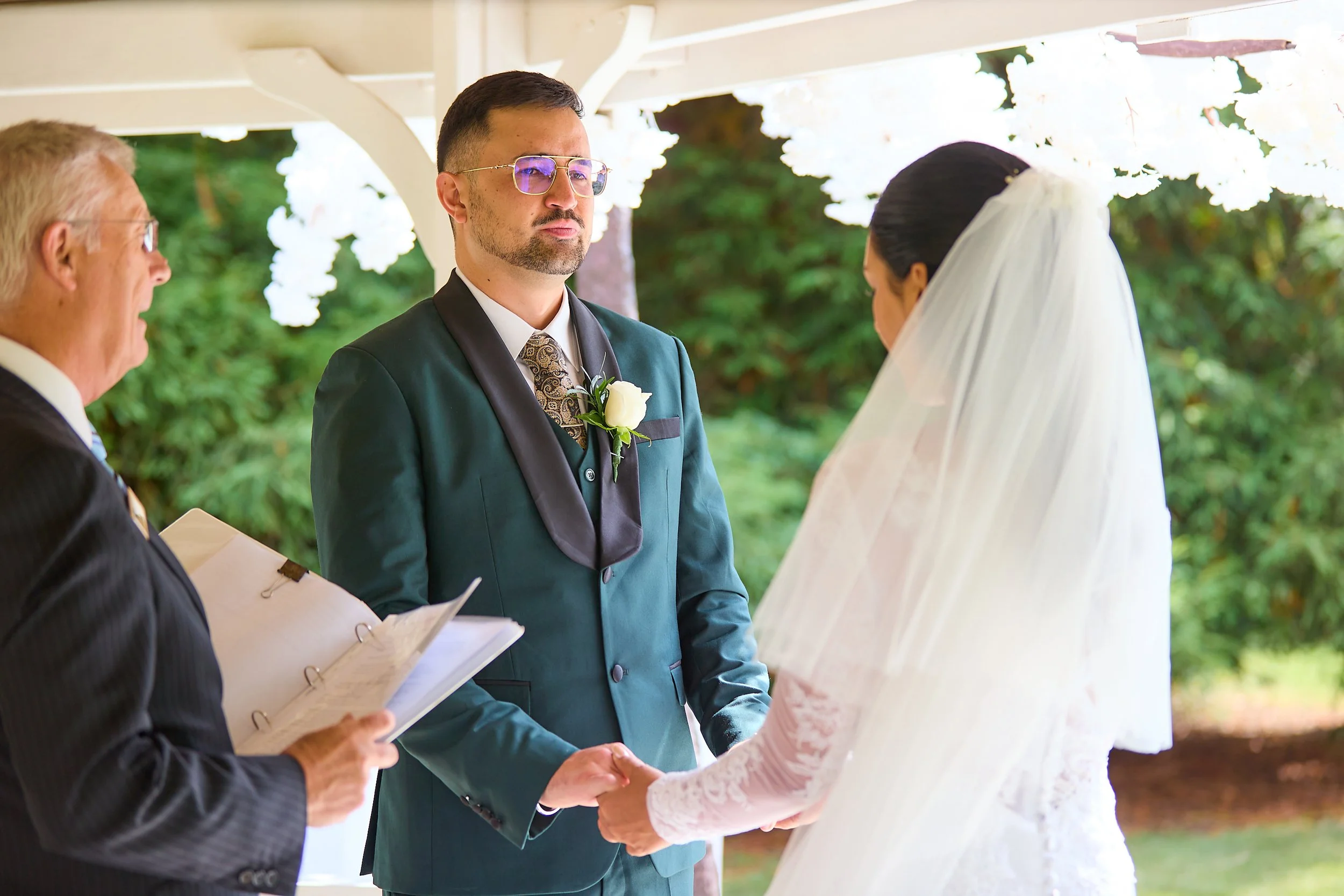 A bride and groom holding hands during their wedding ceremony, with an officiant reading from a book at a decorated outdoor venue.