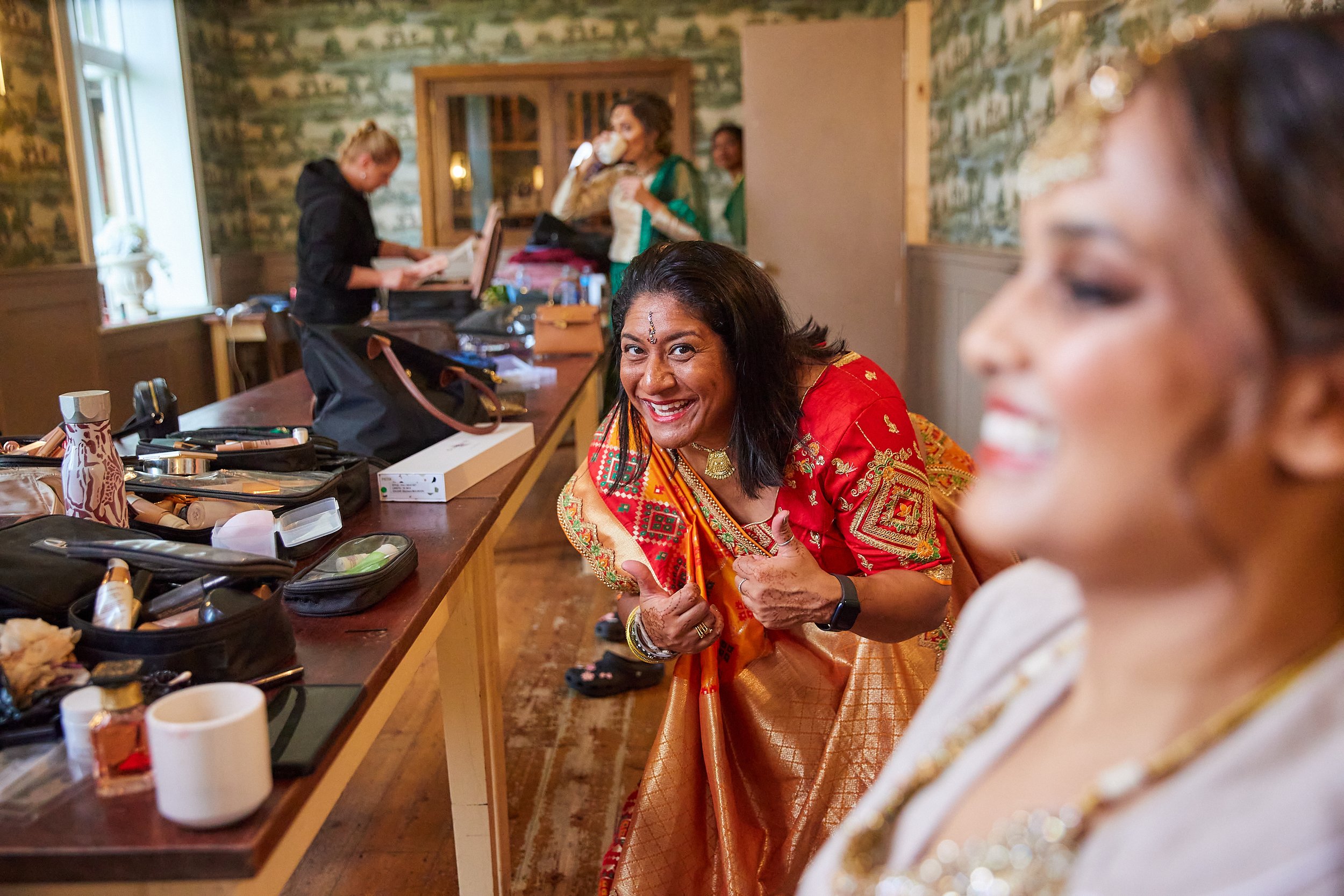 Women getting ready, one smiling and giving a thumbs up, in a room with makeup supplies on the table.