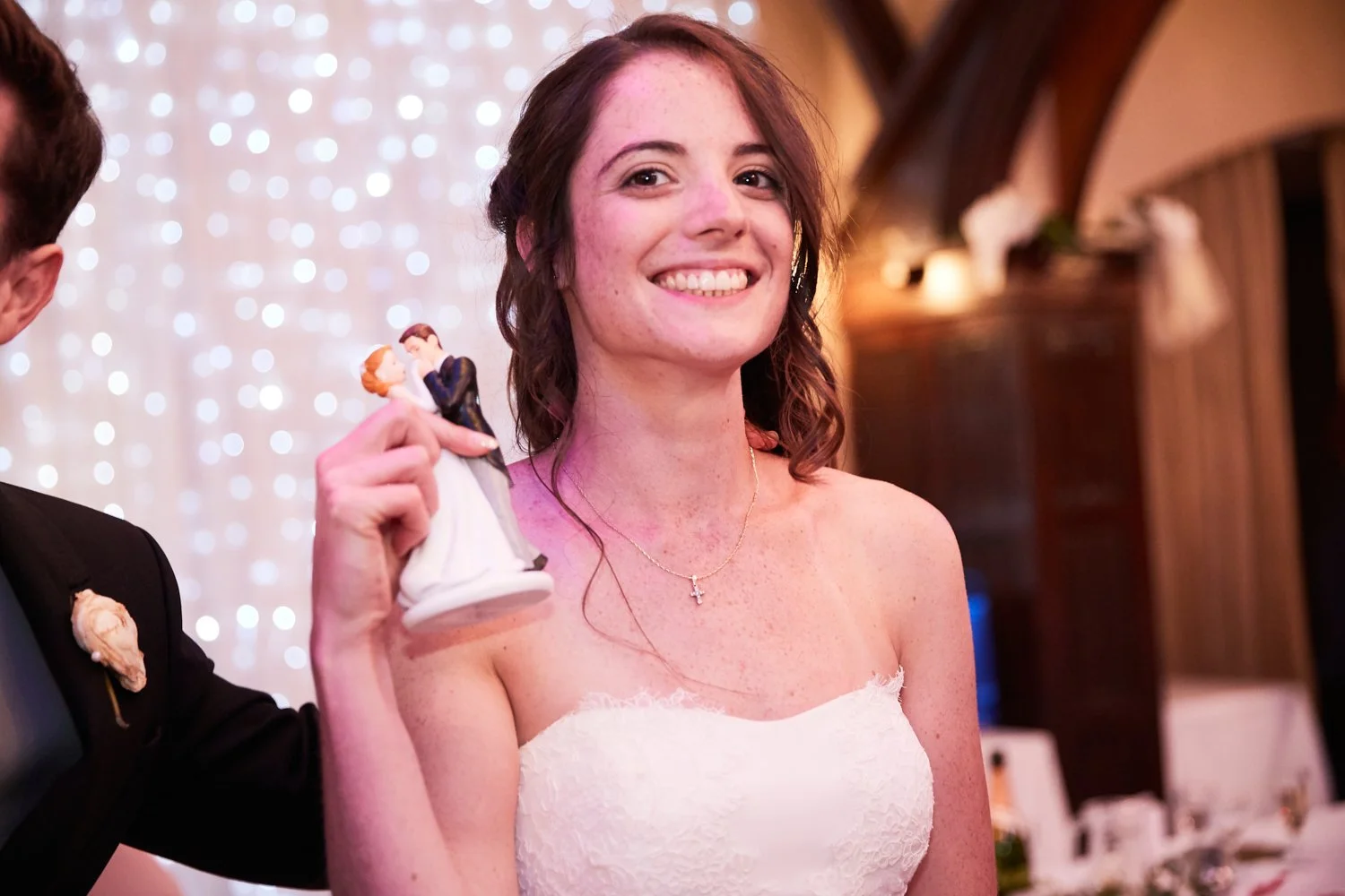 A young woman in a white strapless dress smiling at a wedding reception, holding a wedding cake topper figurine of a bride and groom in her right hand.