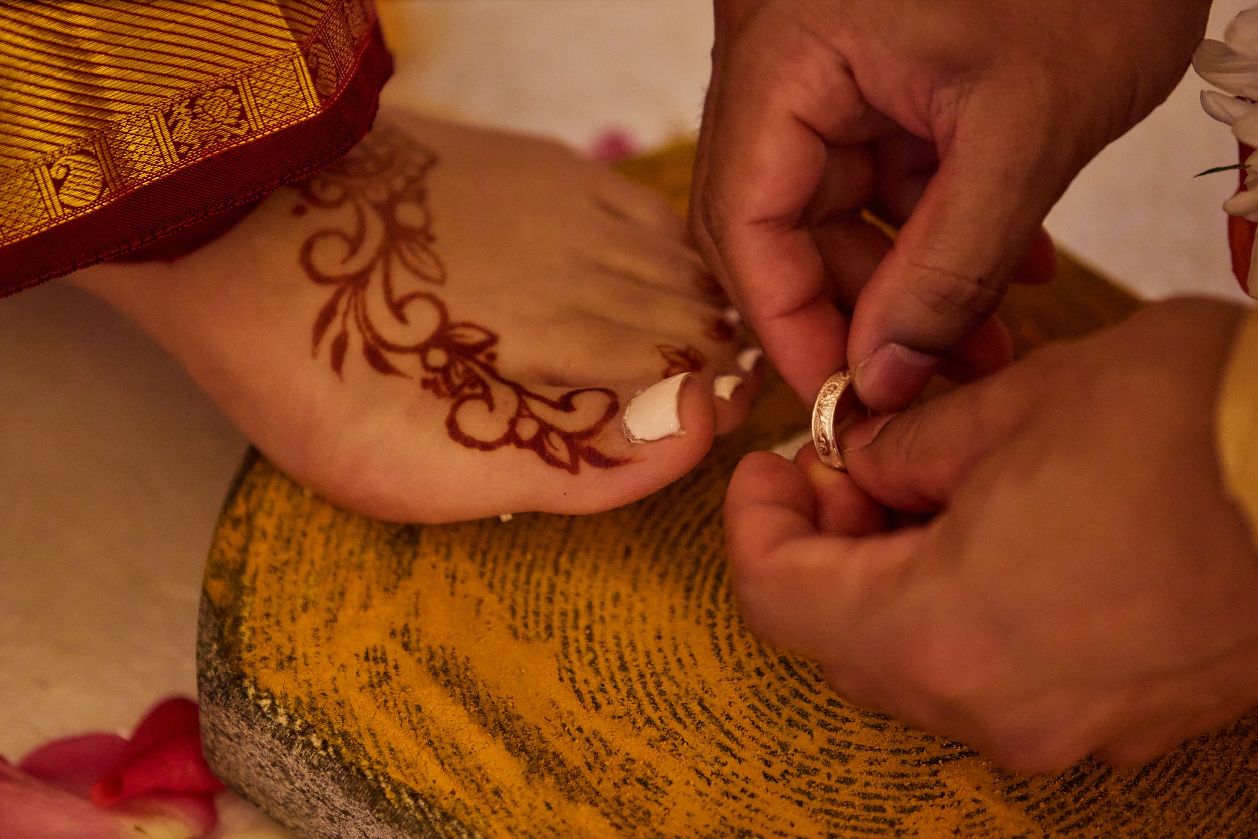 A person wearing traditional Indian attire is placing a ring on another person's hand during a wedding ceremony, with a henna design visible on the hand.