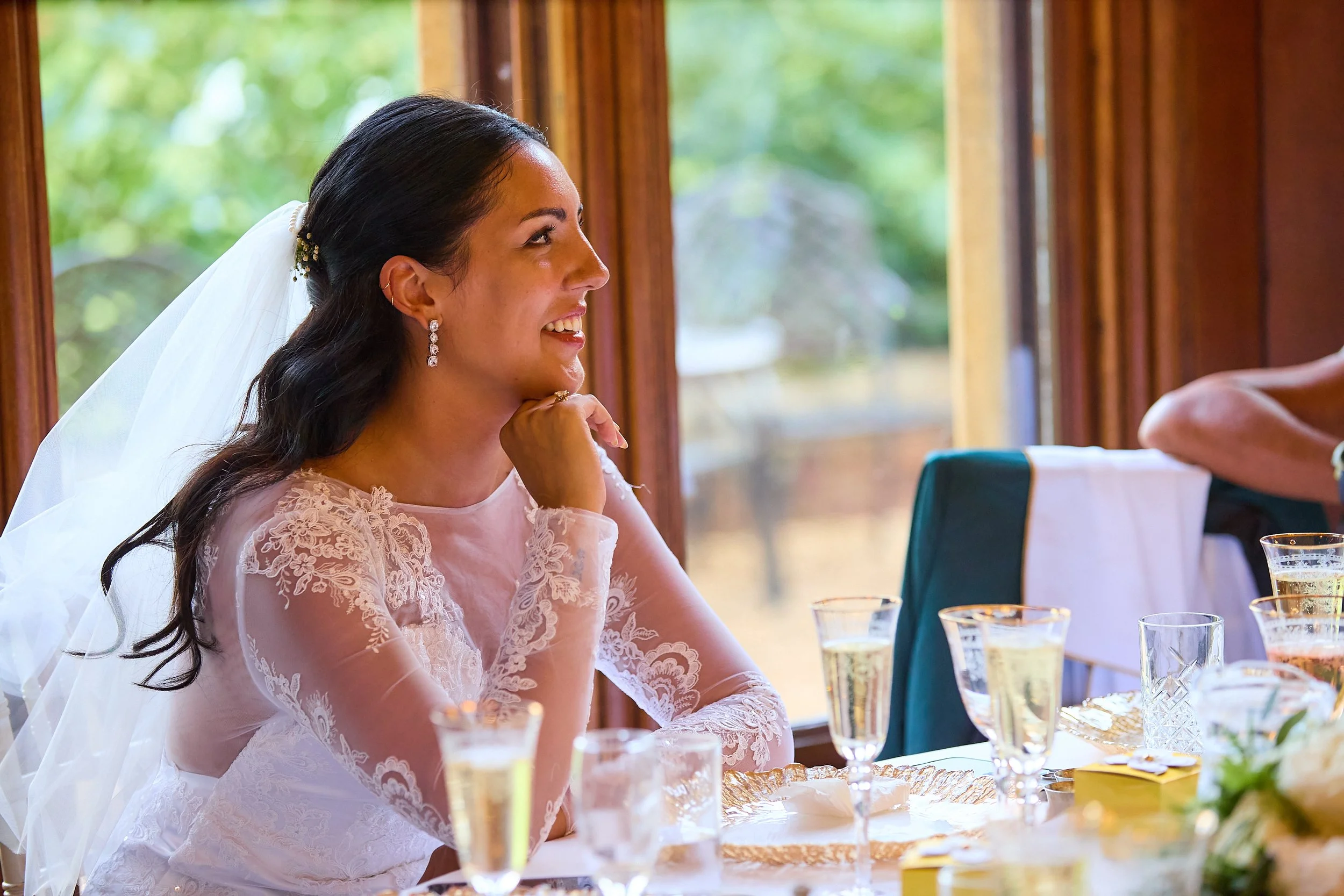 A bride sitting at a table, smiling and looking out a window, with glasses of champagne and floral decorations in front of her.