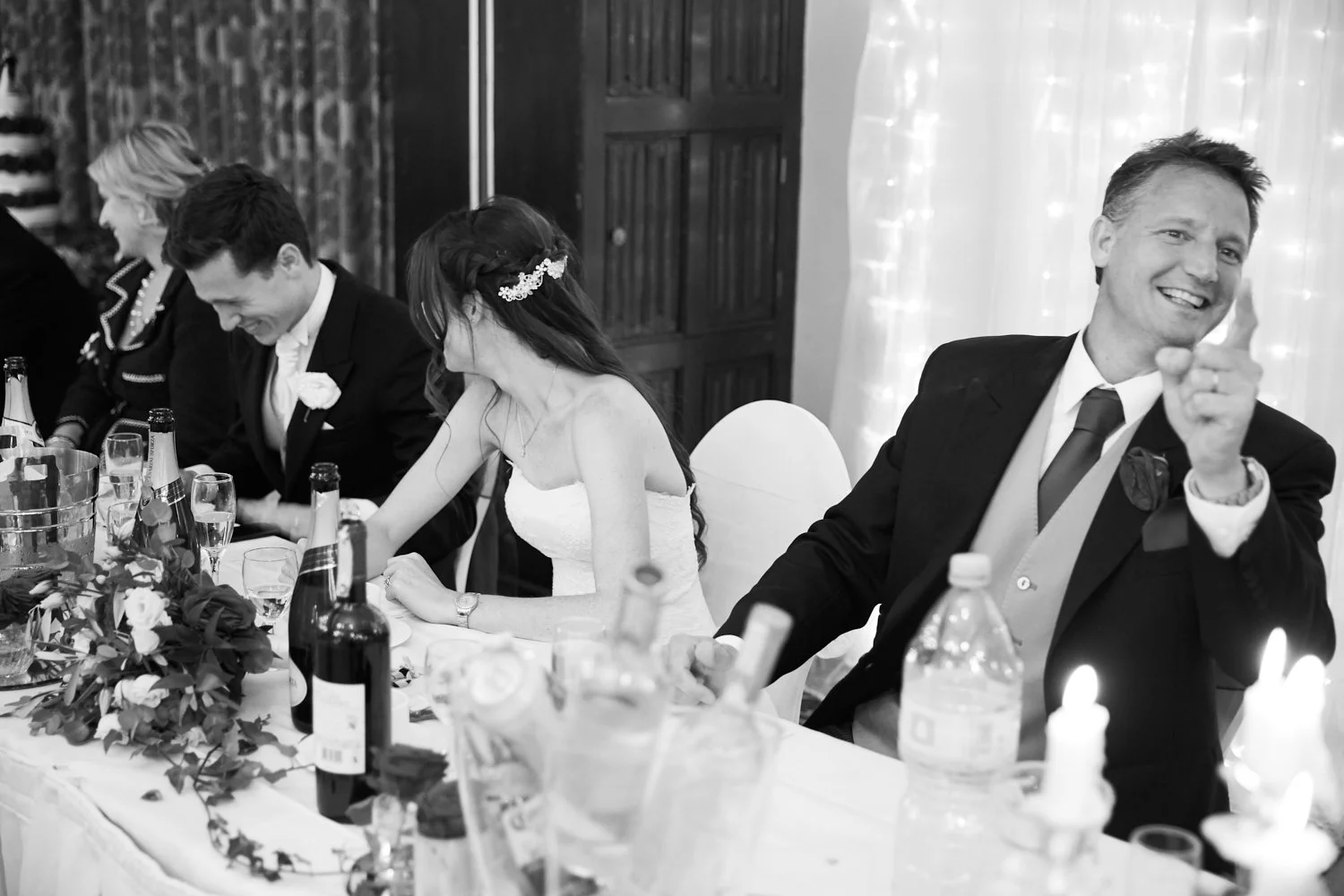 A black and white photo of a wedding reception table with guests, including a bride in a strapless wedding dress, a groom in a suit, and a smiling man pointing, with bottles, glasses, and floral decorations on the table.