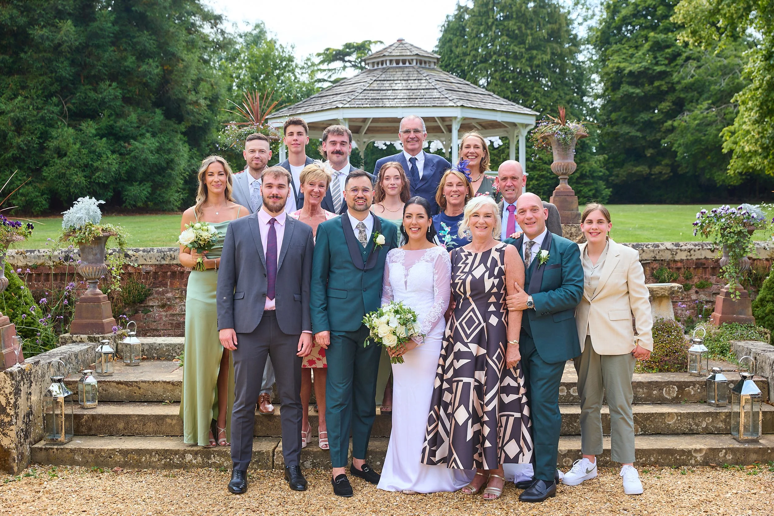 Group of people at a wedding outdoors, standing on stone steps in front of a gazebo, with green trees and grass in the background, some holding bouquets of flowers.