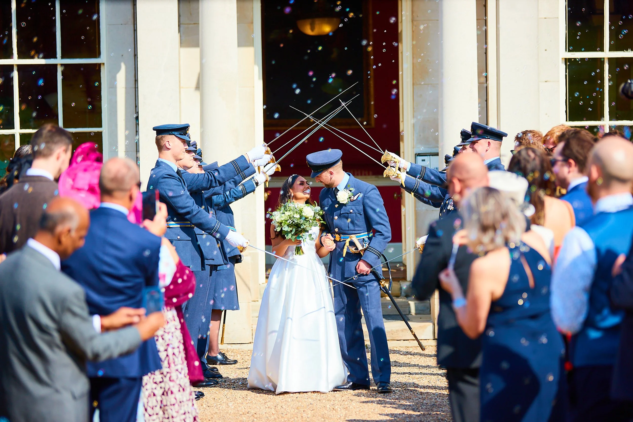 A wedding couple stands in the center surrounded by people outside a building, with soldiers in uniform forming a celebratory arch with swords.