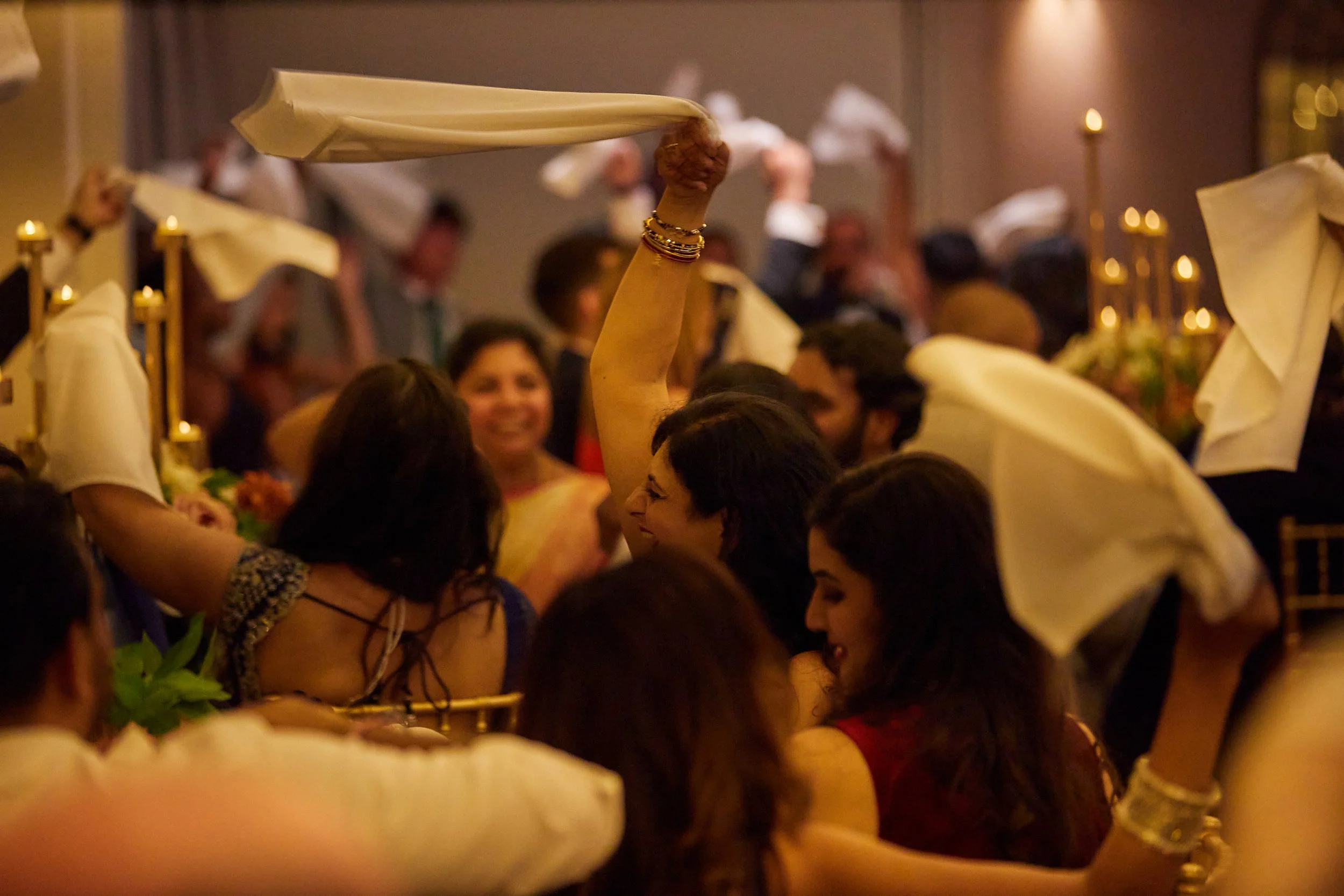 Guests celebrate at a festive gathering with smiles and waving cloth napkins, decorated with candles and flowers in the background.