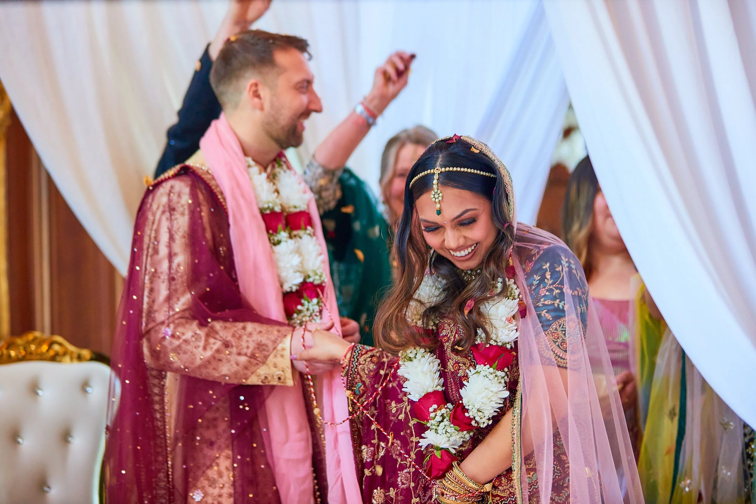 A bride and groom celebrate their wedding, smiling and holding hands under a decorated canopy, with friends and family around.