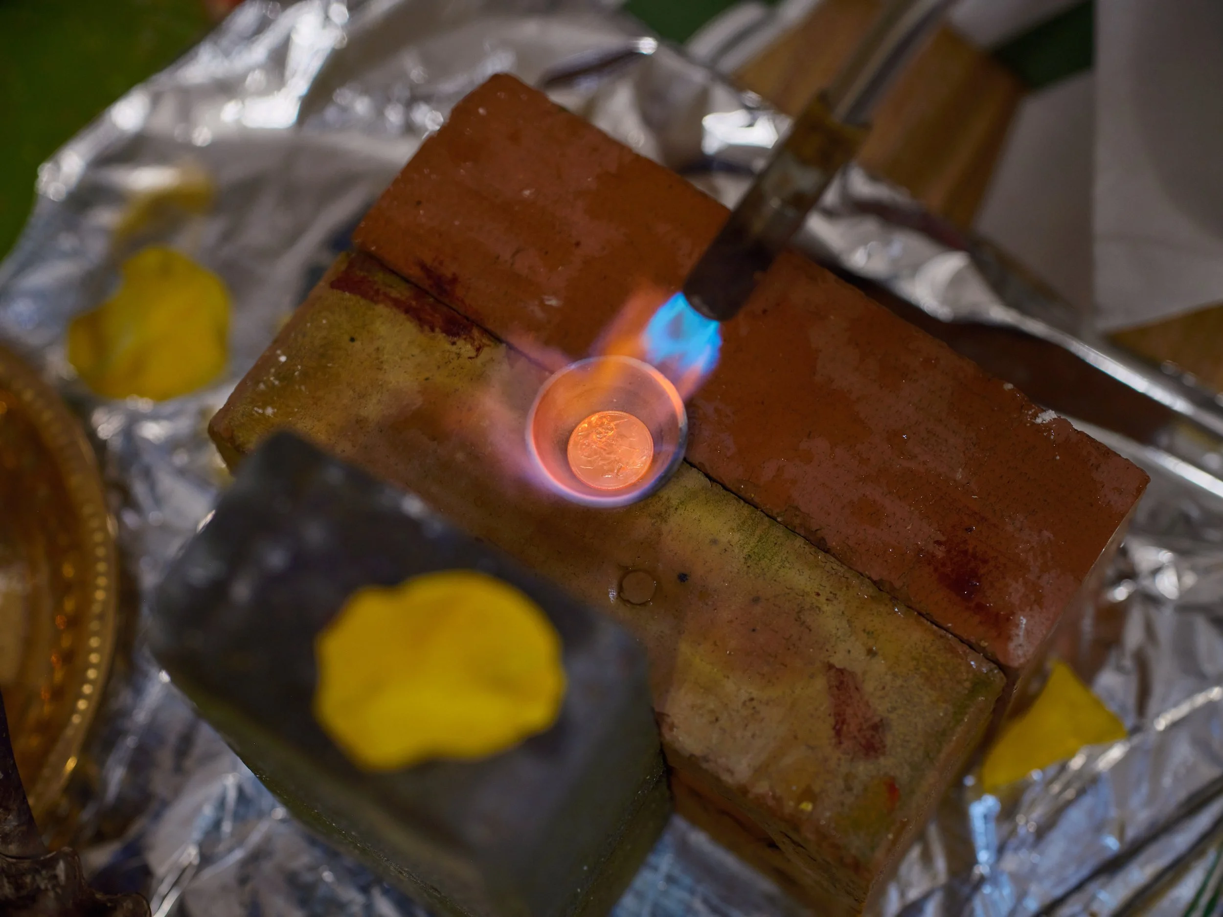Bricks on a foil-wrapped surface with a propane torch heating one brick, with yellow flower petals nearby.