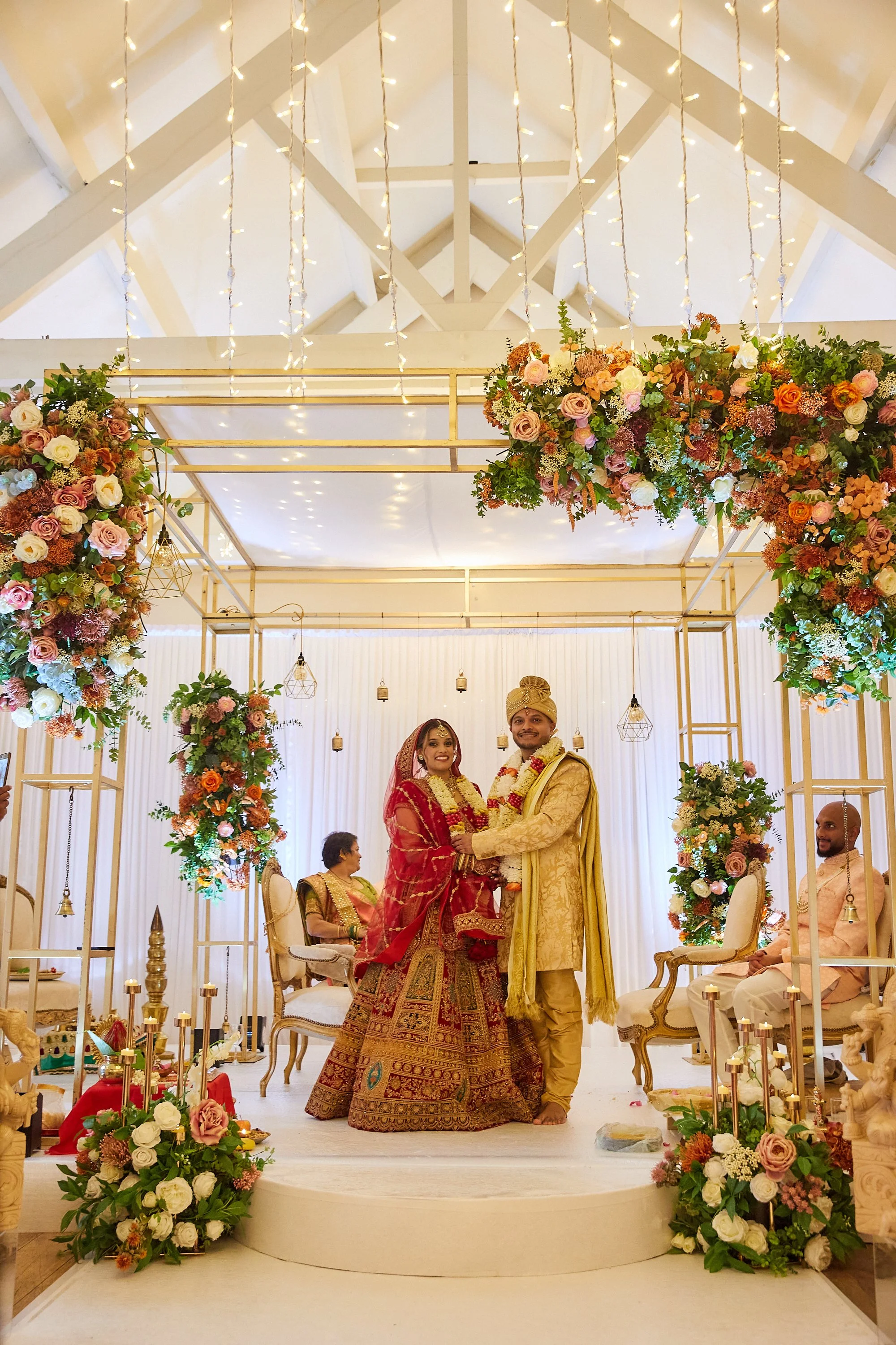 A wedding ceremony with a bride and groom dressed in traditional Indian attire, standing on a decorated stage with floral arrangements and hanging lights, with two seated officiants or family members in the background.