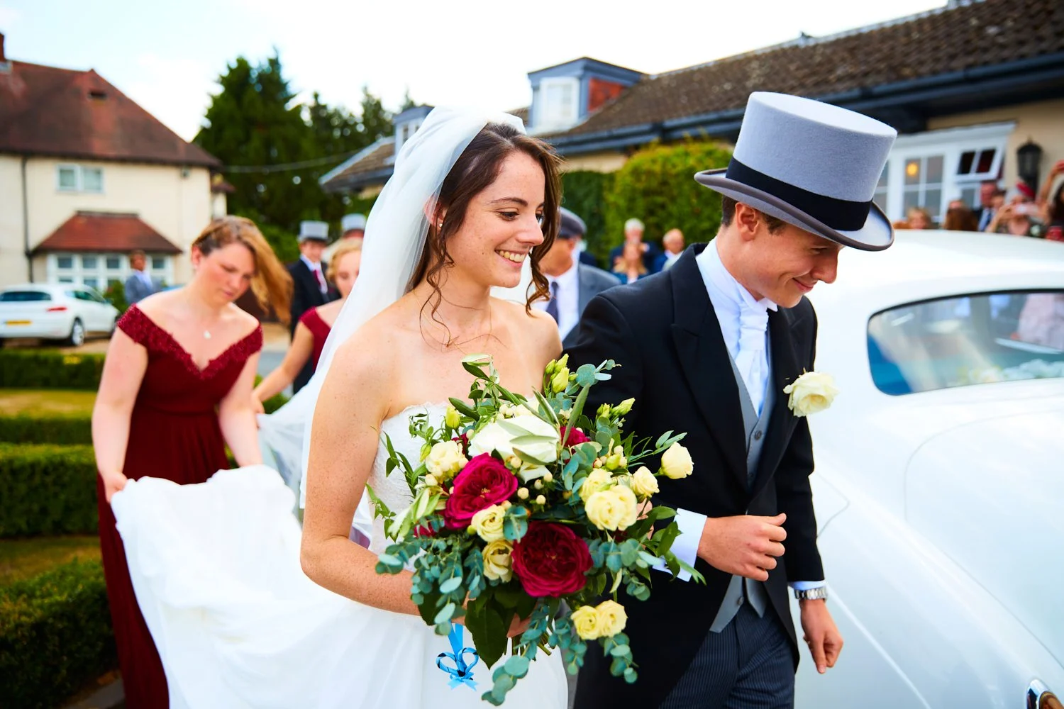 A bride and groom smiling and holding a bouquet of flowers, getting into a white vintage car at their wedding celebration, with guests in the background.
