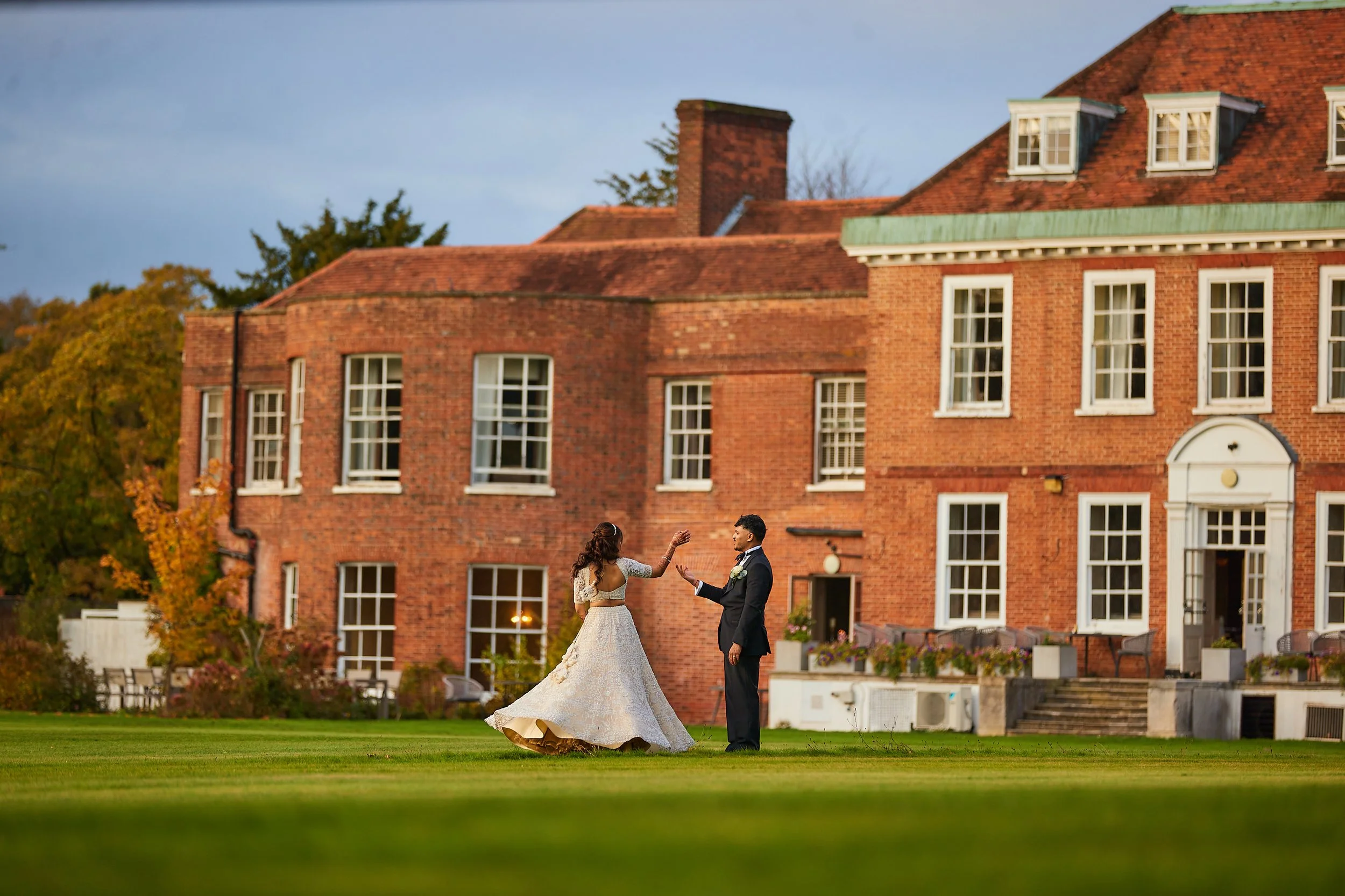 A bride and groom dancing on a green lawn in front of a large red brick house during sunset.