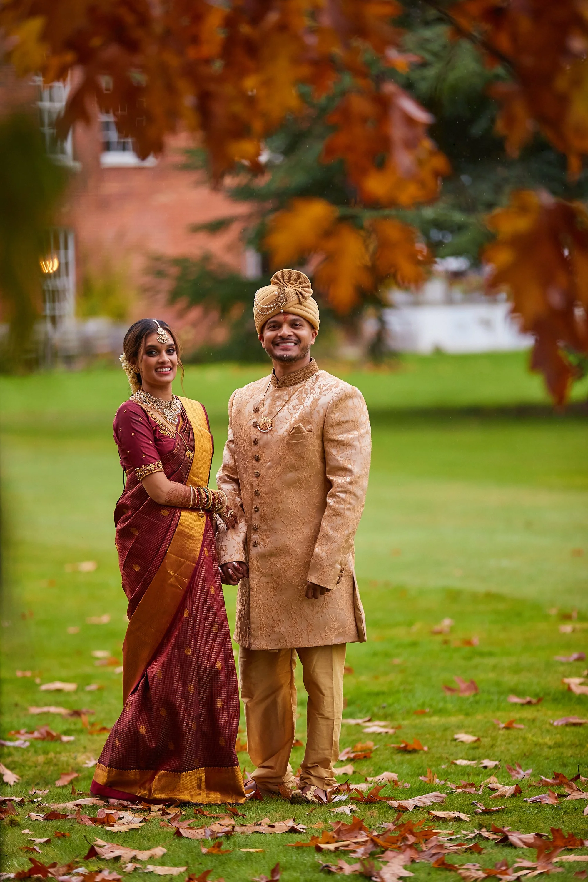 A couple in traditional Indian wedding attire standing outdoors on a grassy area with fallen leaves, trees, and a building in the background. The woman wears a maroon and gold saree with jewelry, and the man is dressed in a beige and gold sherwani wi