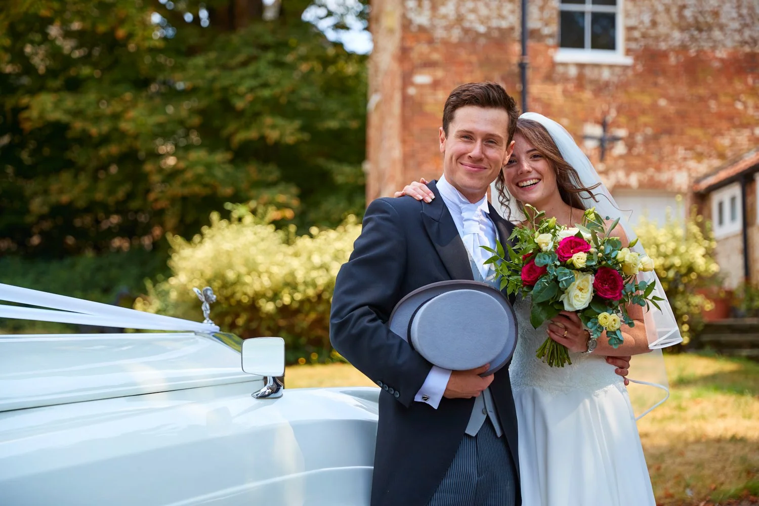 A newly married couple standing outdoors near a white vintage car, smiling and embracing. The groom is in a dark suit holding a gray bowler hat, and the bride is in a white wedding dress holding a colorful bouquet of flowers. There are trees and a br