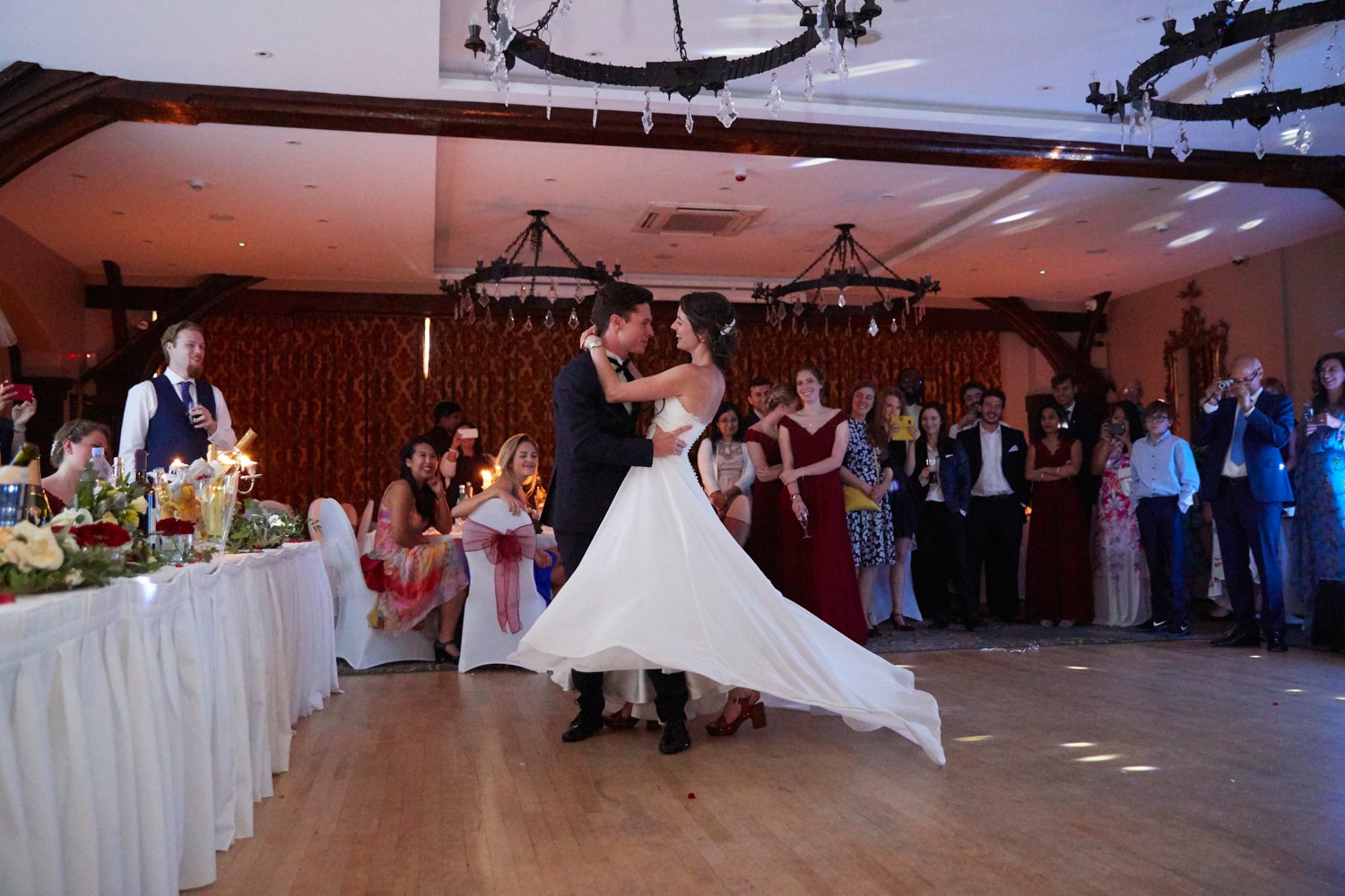 A bride and groom are dancing in the center of a wedding reception with guests observing and taking photos in the background. The bride is wearing a white wedding gown, and the groom is in a black tuxedo. The guests are seated and standing around the