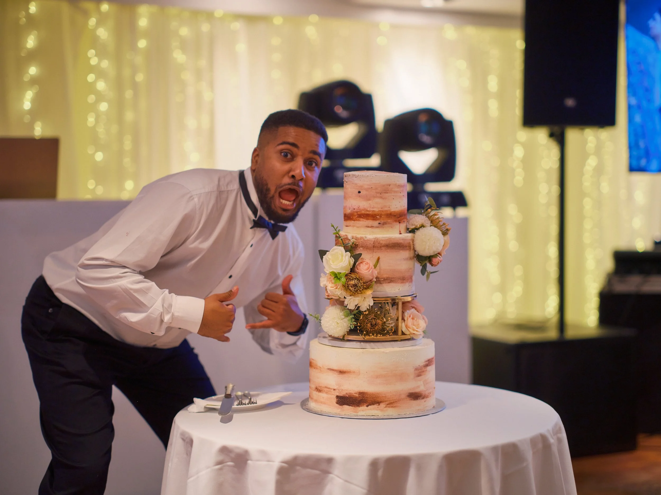 A man dressed as a waiter at a wedding reception giving a thumbs up and making a surprised face next to a three-tiered wedding cake decorated with flowers and greenery.
