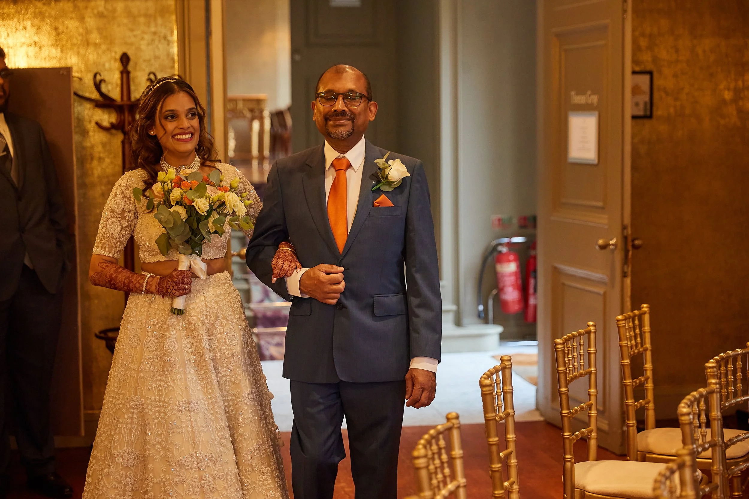 A bride in a lace wedding dress holding a bouquet of flowers and an older man in a suit with an orange tie and boutonniere walking together at a wedding venue.