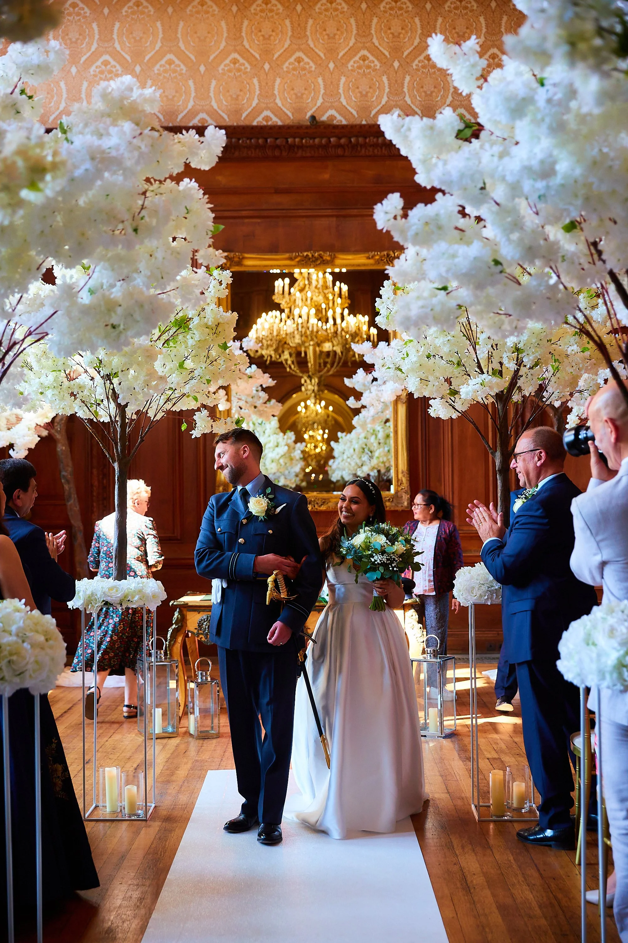 A bride and groom walk through an aisle decorated with white flowers and candles in lanterns, surrounded by guests at a wedding ceremony in a grand, wood-paneled room with chandeliers.