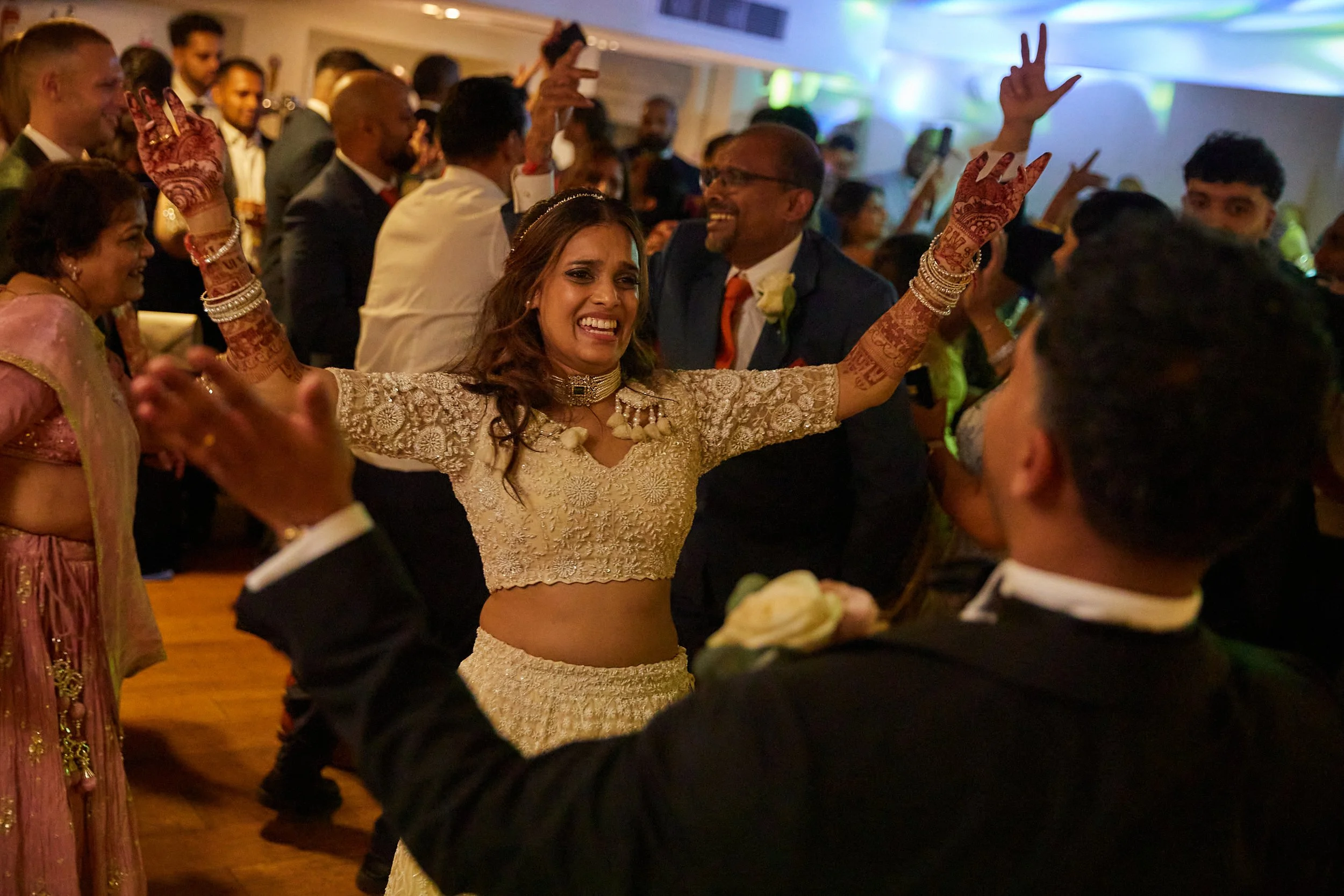 A bride in traditional Indian attire celebrating with guests at her wedding reception, dancing with her arms raised, surrounded by guests in formal clothing at a lively indoor event.