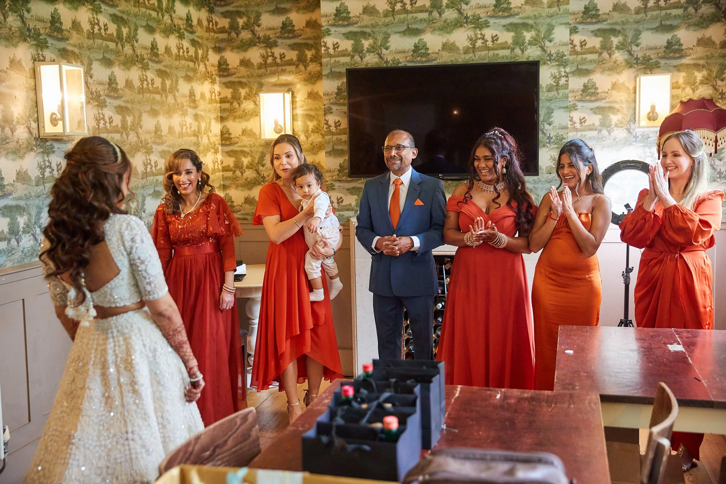 Group of people at a wedding reception, with seven women and one man standing and smiling across from a bride dressed in a white gown. The women wear various shades of orange or red dresses, and the man is in a dark blue suit with an orange tie. The 