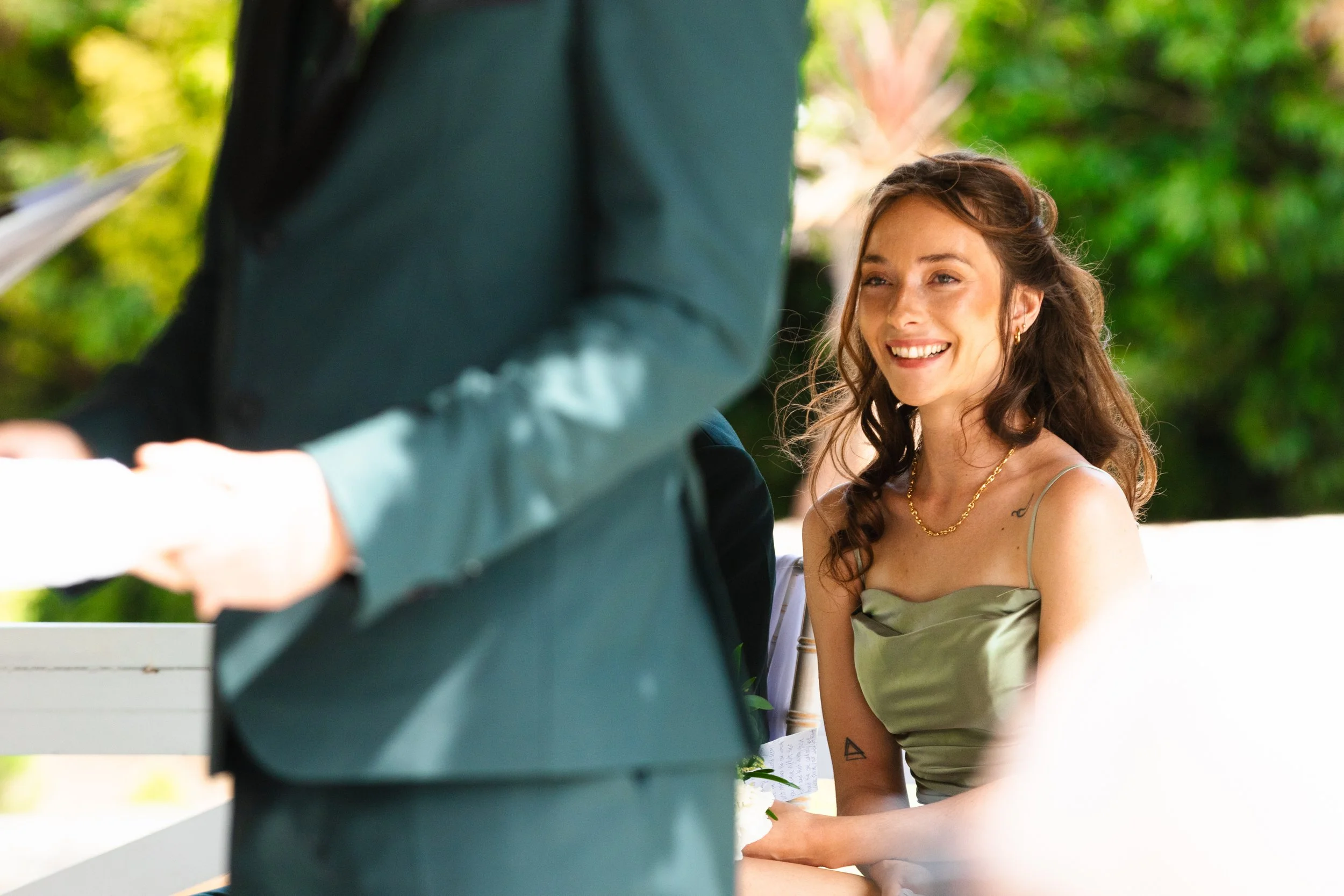 A smiling woman with brown hair wearing a green slip dress, sitting outdoors near greenery, facing a person in a dark suit, with gift boxes in the foreground.
