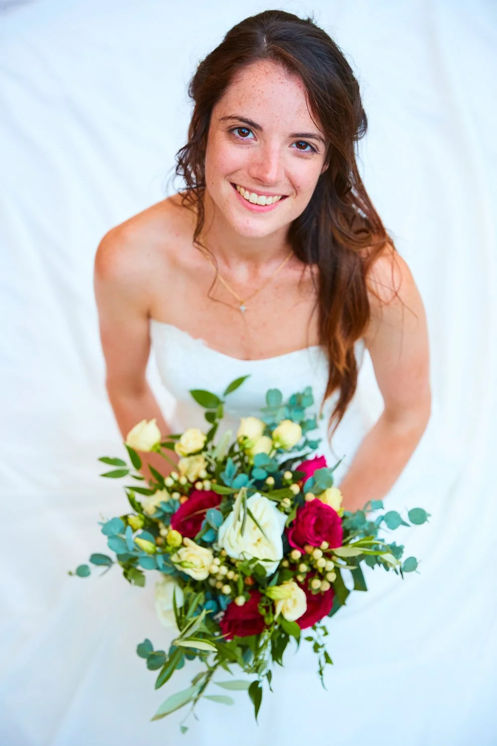 A smiling young woman in a strapless white dress holding a bouquet of flowers, looking up at the camera.