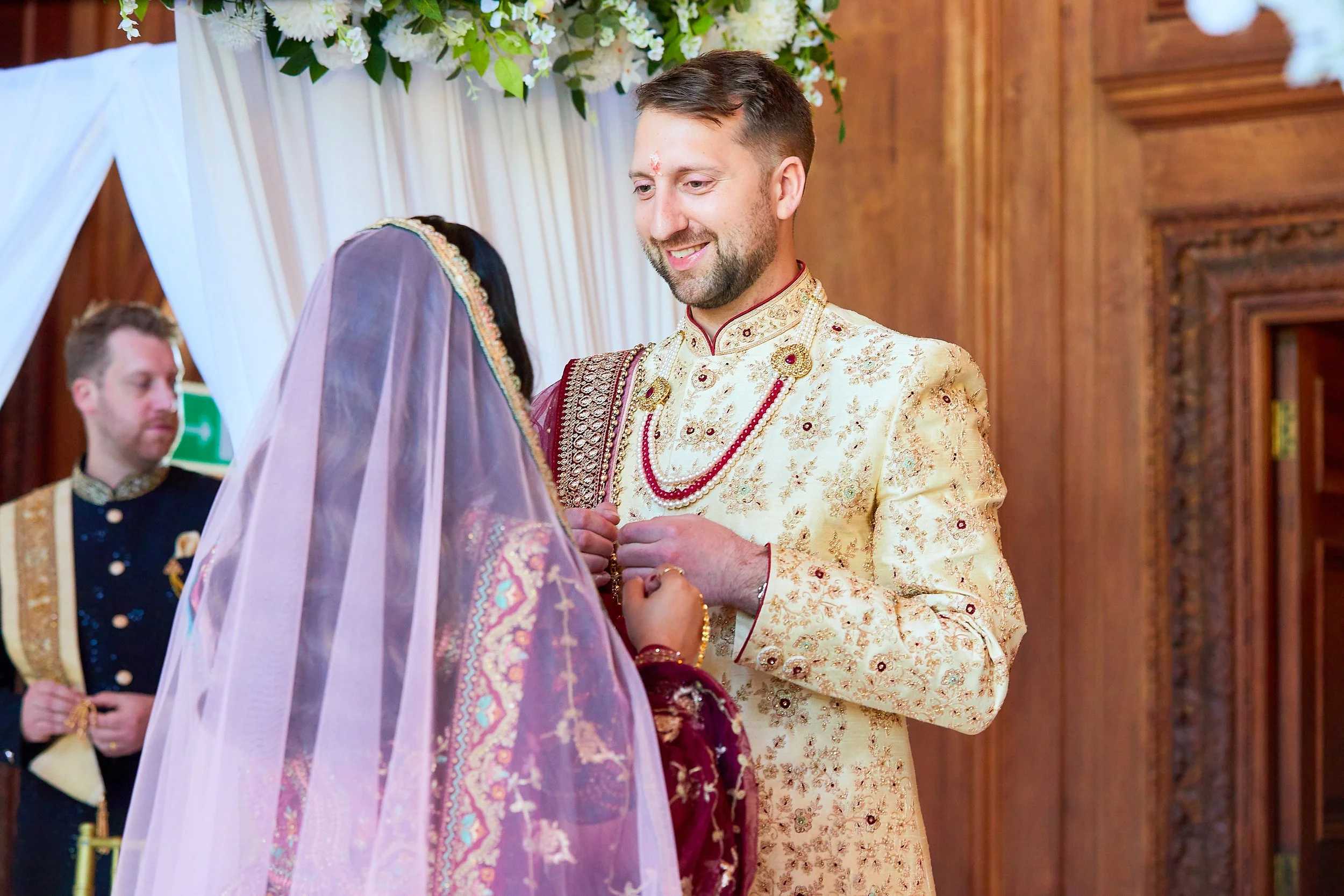 A man and a woman dressed in traditional Indian wedding attire during a ceremony, with the man smiling and holding the woman's hands, a woman with a sari and veil, in a decorated indoor setting.