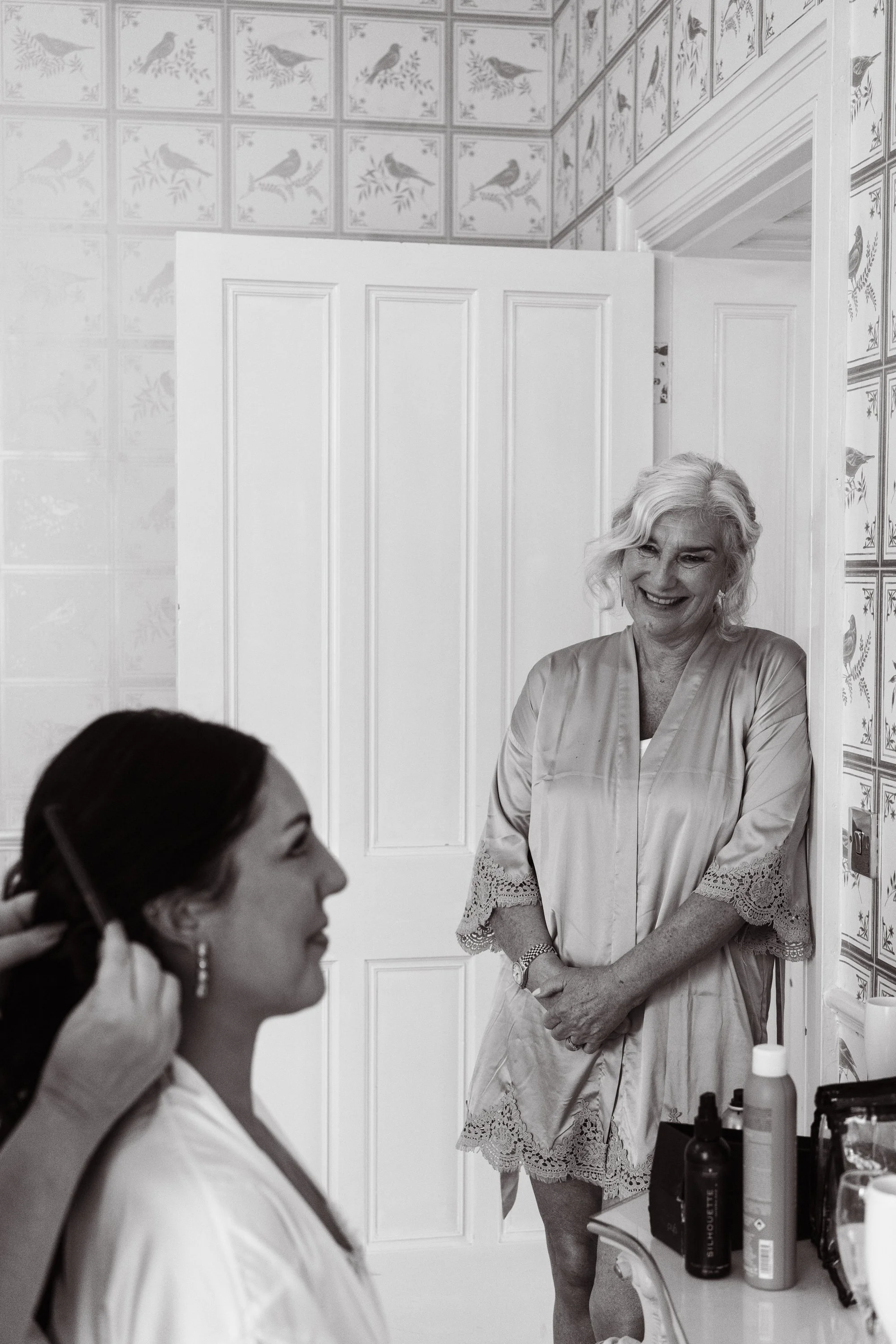 A woman getting her makeup done by another woman in a room with bird-patterned wallpaper, smiling and looking at each other.