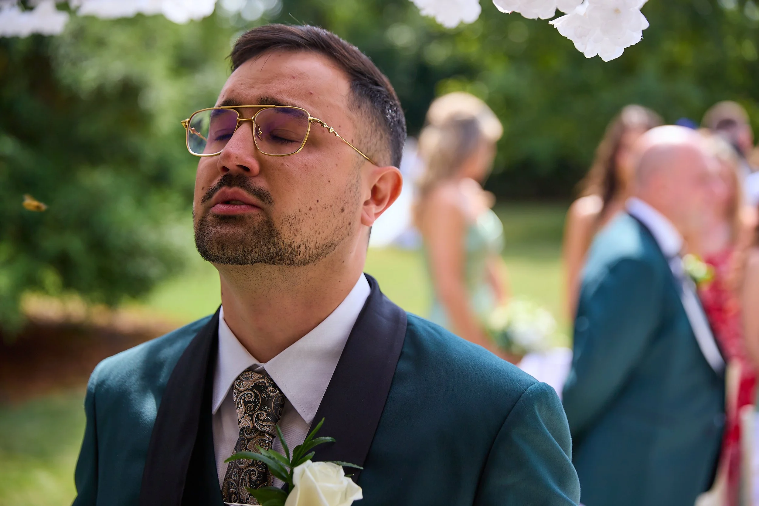 A young man with glasses, wearing a teal suit, white shirt, and patterned tie, has a white rose boutonniere on his lapel. His eyes are closed, and he appears emotional. There are blurred people and greenery in the background, suggesting an outdoor we