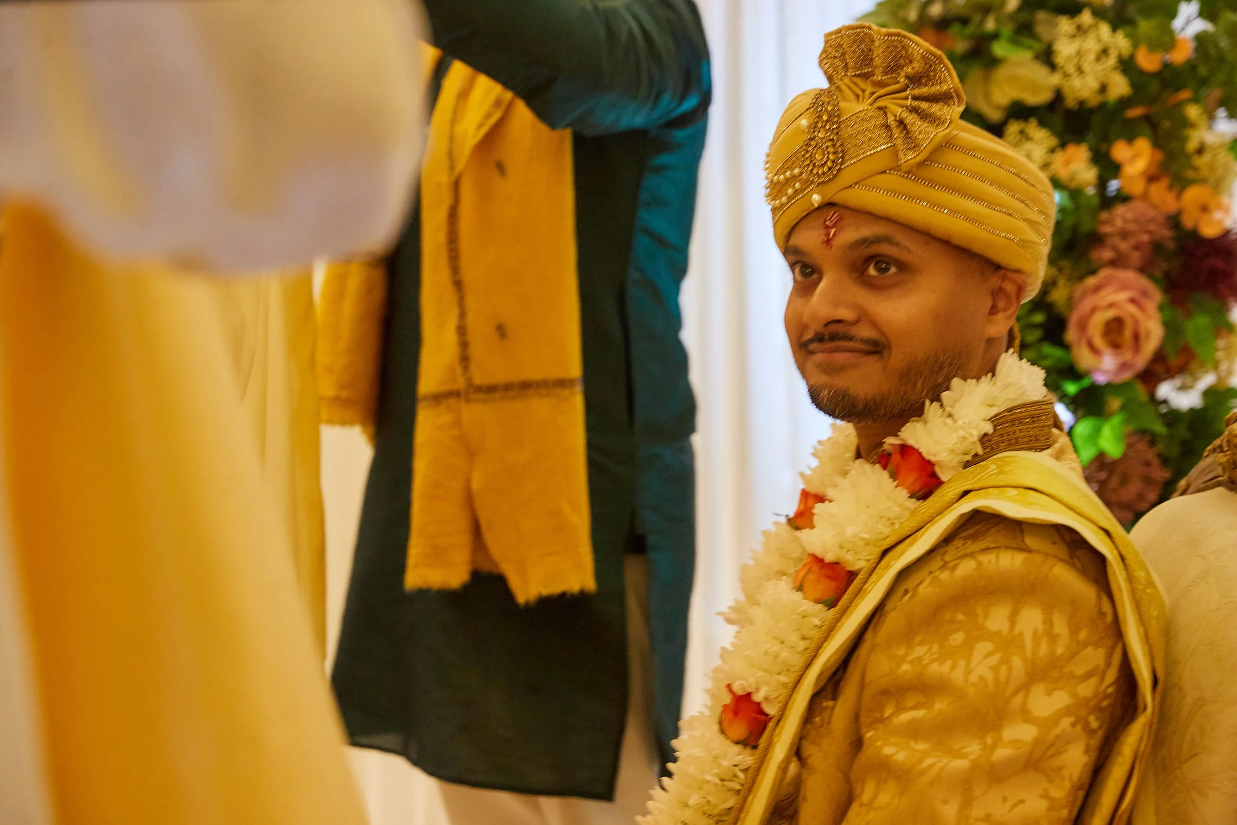 Groom wearing traditional Indian wedding attire, including a yellow turban and sherwani, adorned with a flower garland, seated during a wedding ceremony.