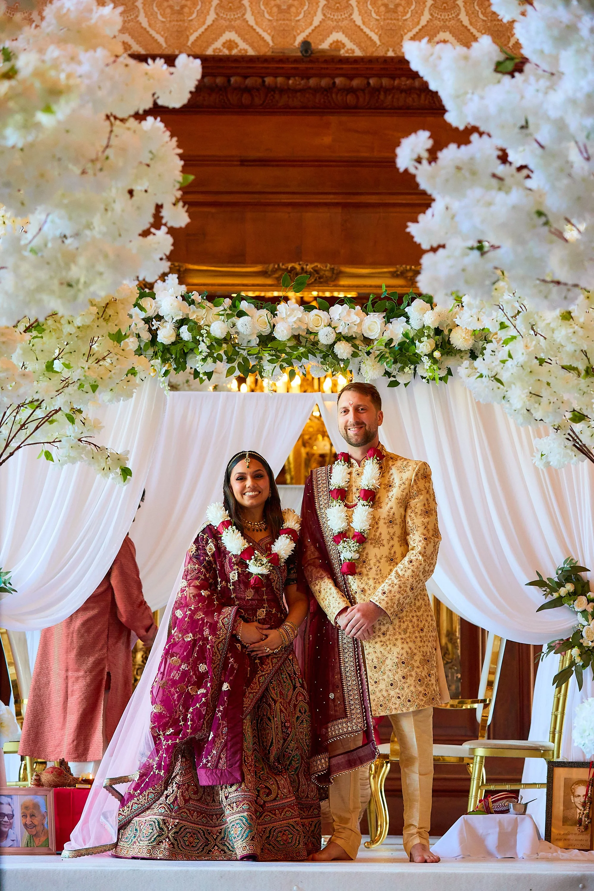A bride and groom standing under a decorated wedding arch, wearing traditional Indian wedding attire, smiling and adorned with flower garlands, in an ornate decorated setting.