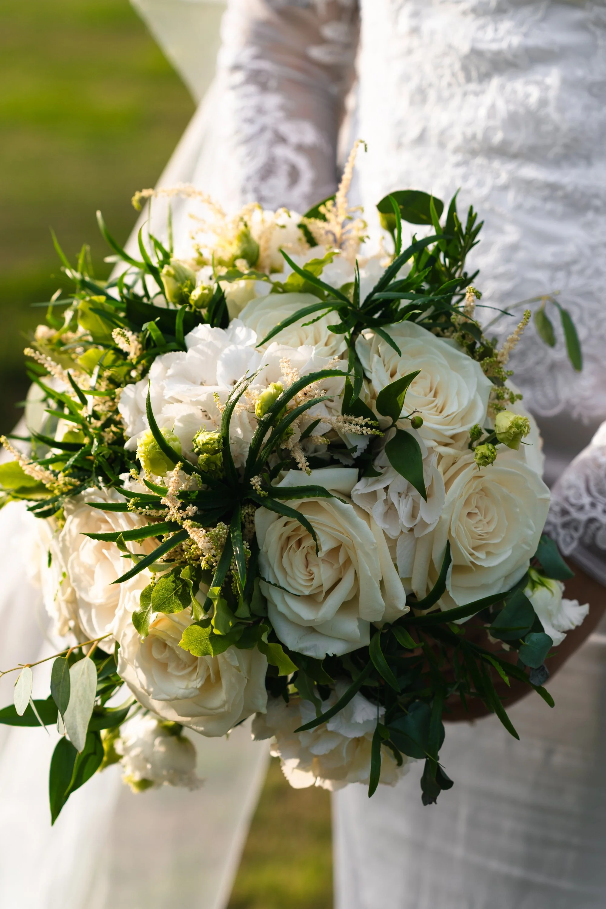 A bride holding a bouquet of white roses and greenery, with a lace wedding dress visible.