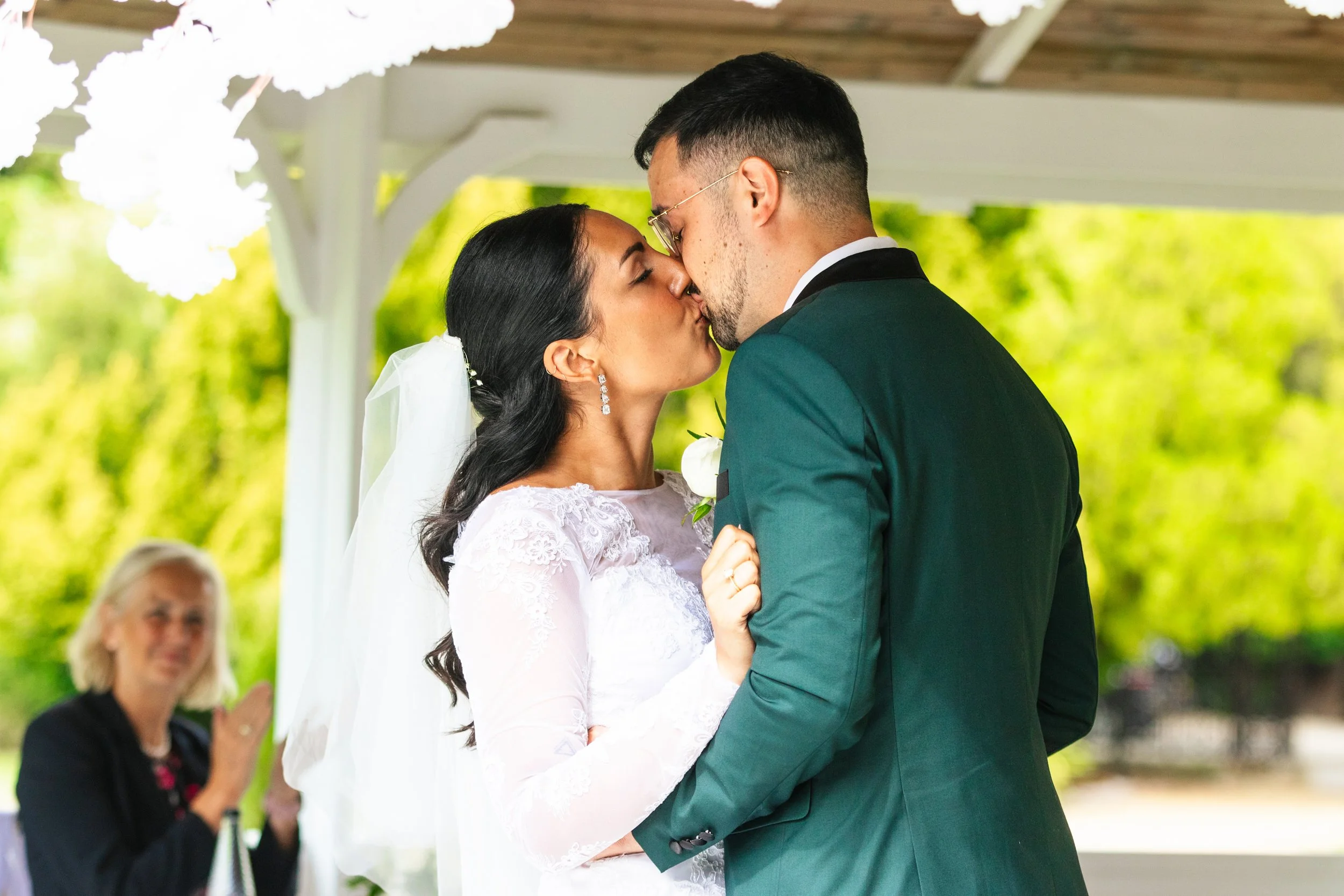 A bride and groom are kissing during their wedding ceremony under a white pavilion, with a woman clapping in the background.