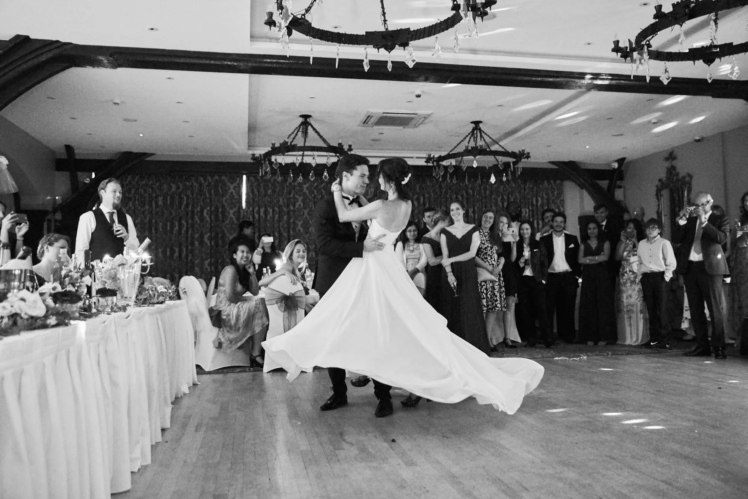 Black and white photo of a wedding reception with a bride and groom dancing in the center, surrounded by guests watching and taking photos, with tables decorated with flowers and a chandelier hanging from the ceiling.
