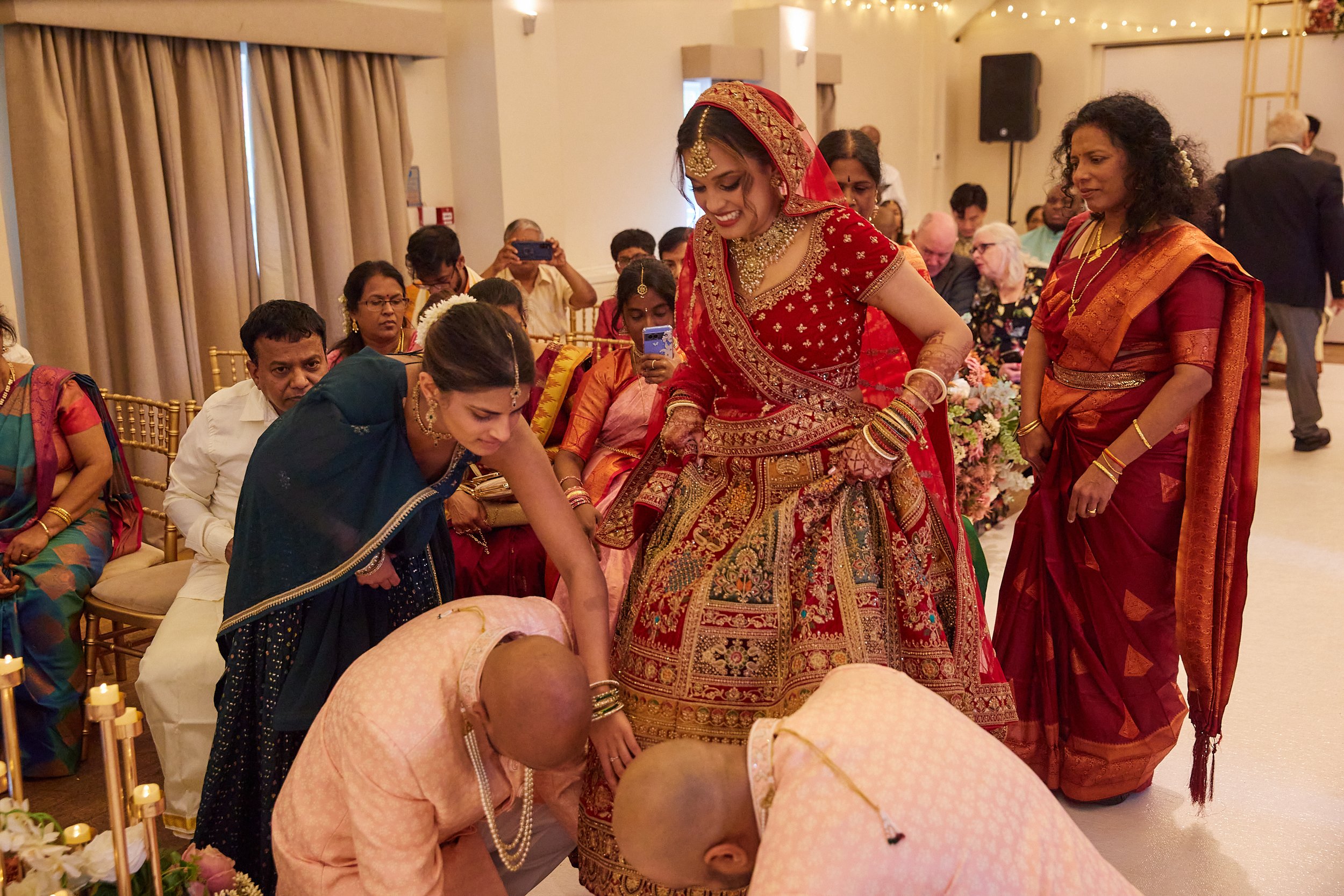 Indian bride dressed in red and gold lehenga with jewelry and henna, participating in a traditional wedding ritual. Surrounded by family and guests, some are bowing to the bride, in a decorated indoor venue.