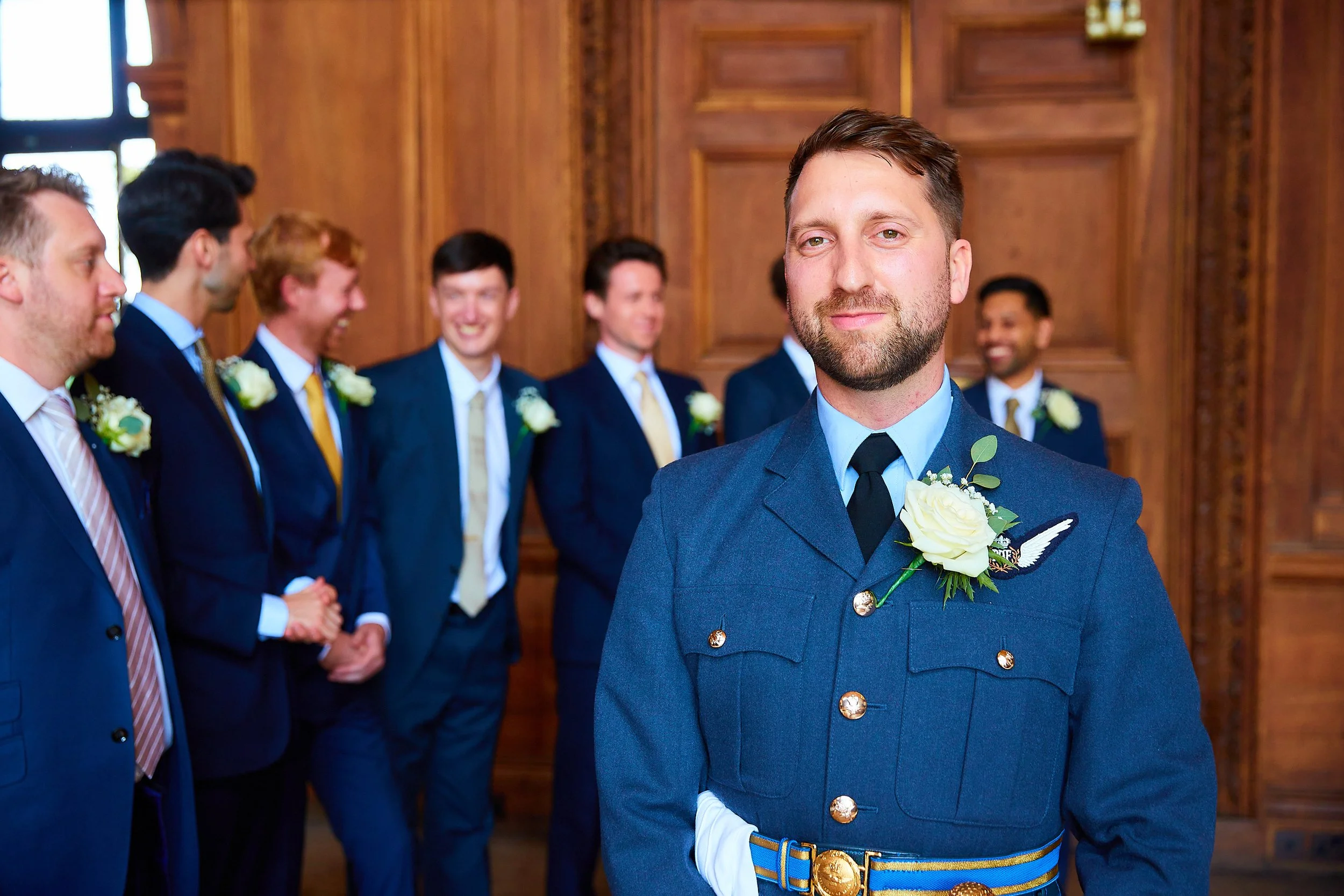 Man in military uniform with boutonnière, standing in front of a group of well-dressed men at a formal event or wedding in a wood-paneled room.