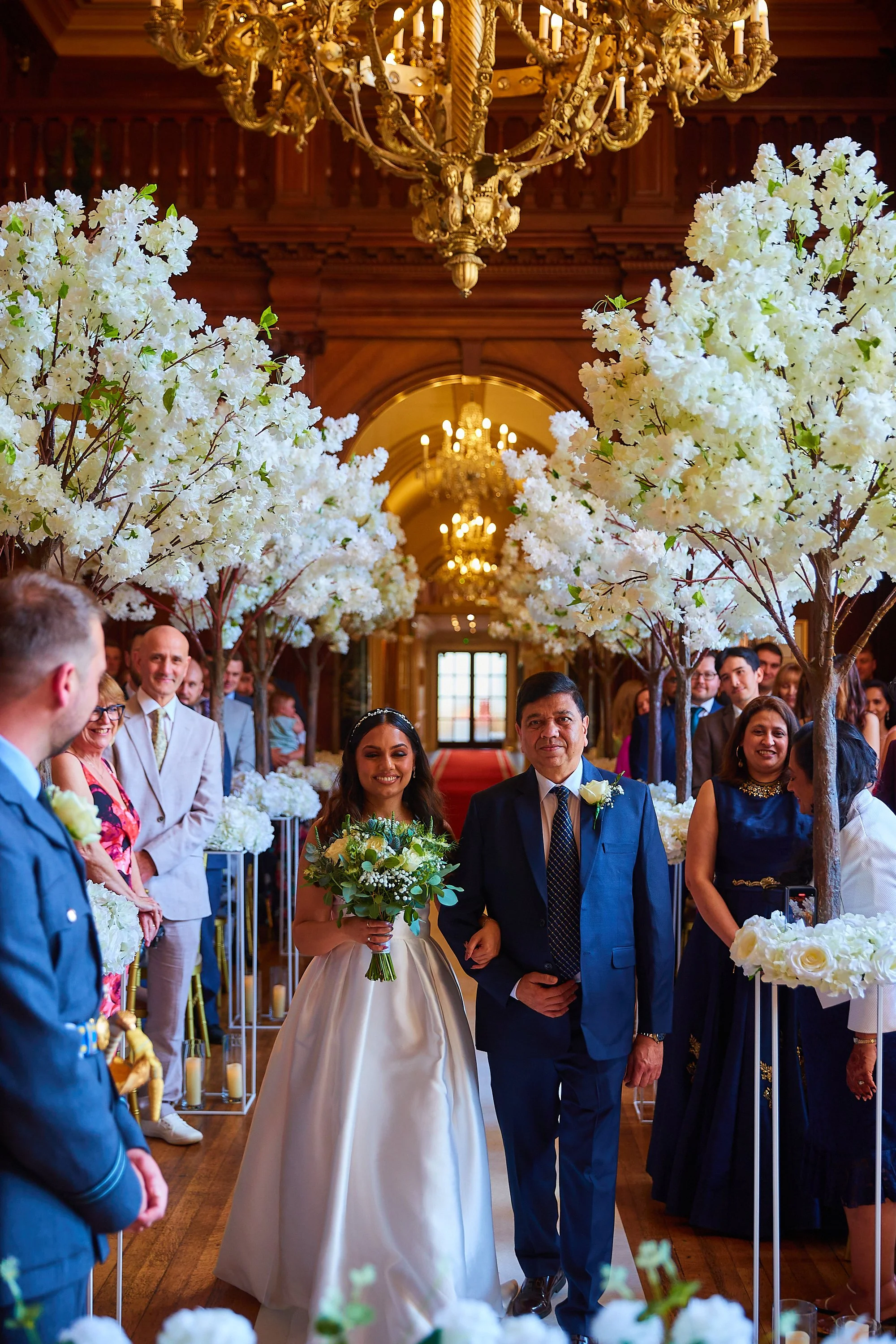A bride, holding a bouquet, walks down the aisle with her father at a wedding ceremony inside a grand, ornate hall. The aisle is decorated with tall white-flowered trees, and guests are smiling and watching the procession.