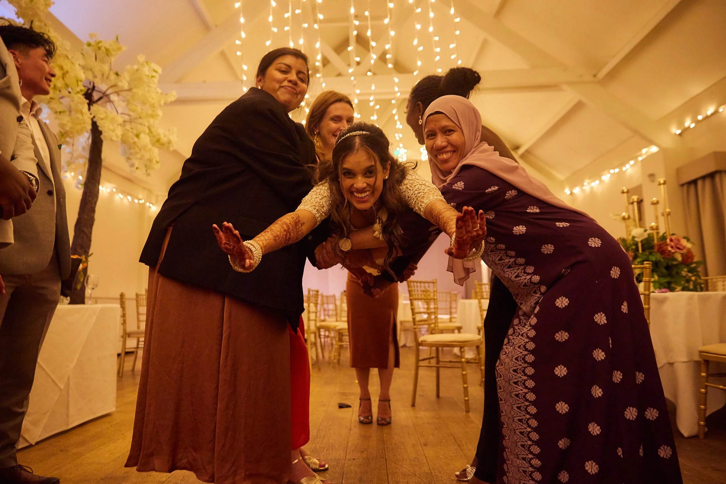 People smiling and posing for a photo at a celebration or party, with decorative lighting and floral arrangements in the background.