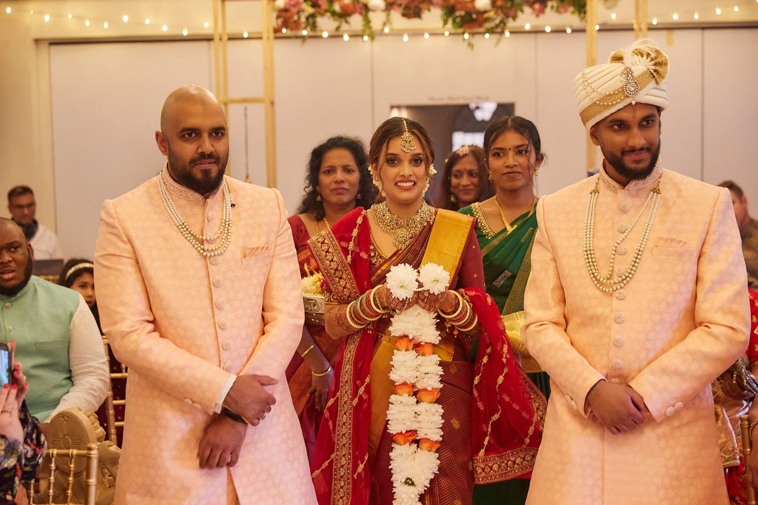 A group of people at a traditional Indian wedding ceremony, with the bride in a red saree and gold jewelry, holding a floral garland, and two men in peach-colored kurtas standing on either side of her, all indoors with warm lighting and decorations.