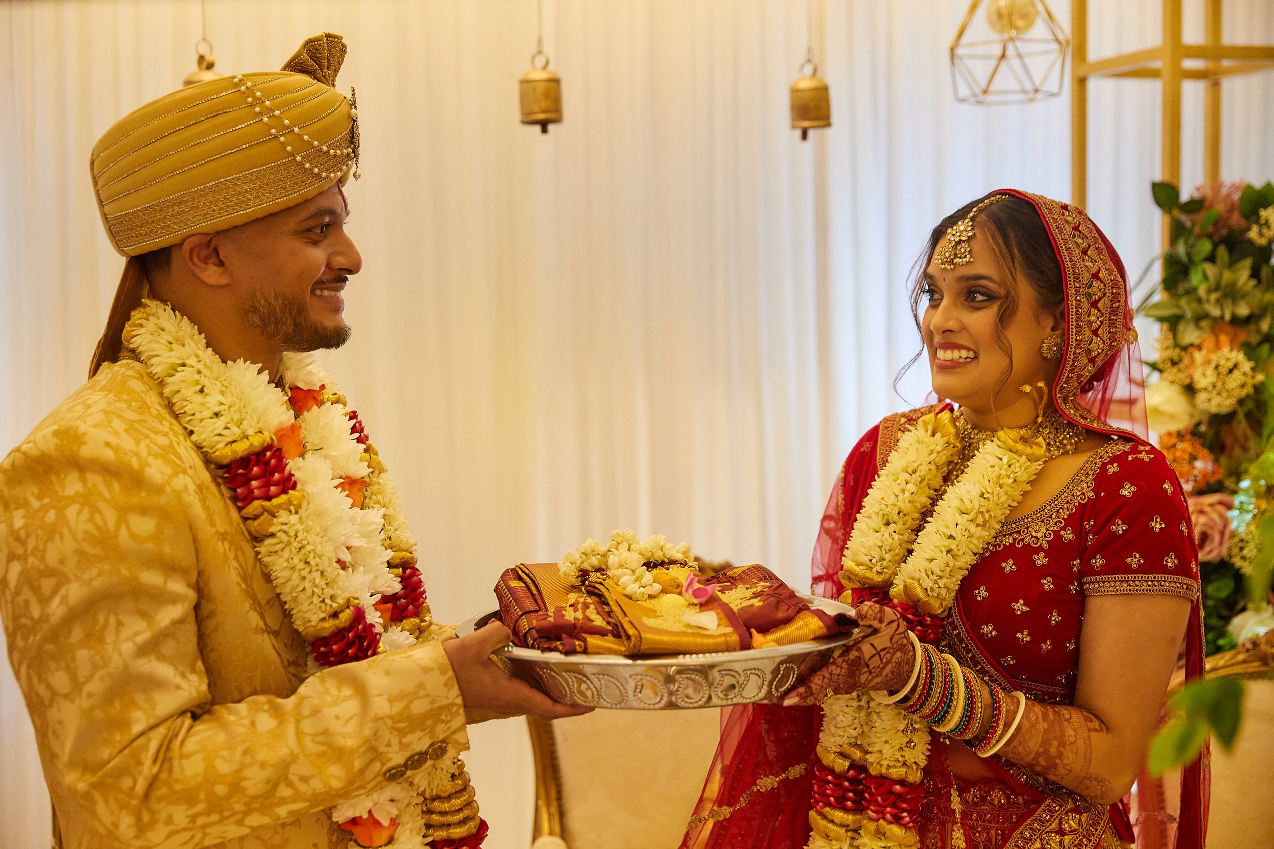 Indian bride and groom during wedding ceremony, exchanging garlands, couple smiling, decorated with flowers, traditional attire, indoors with lights and flowers.