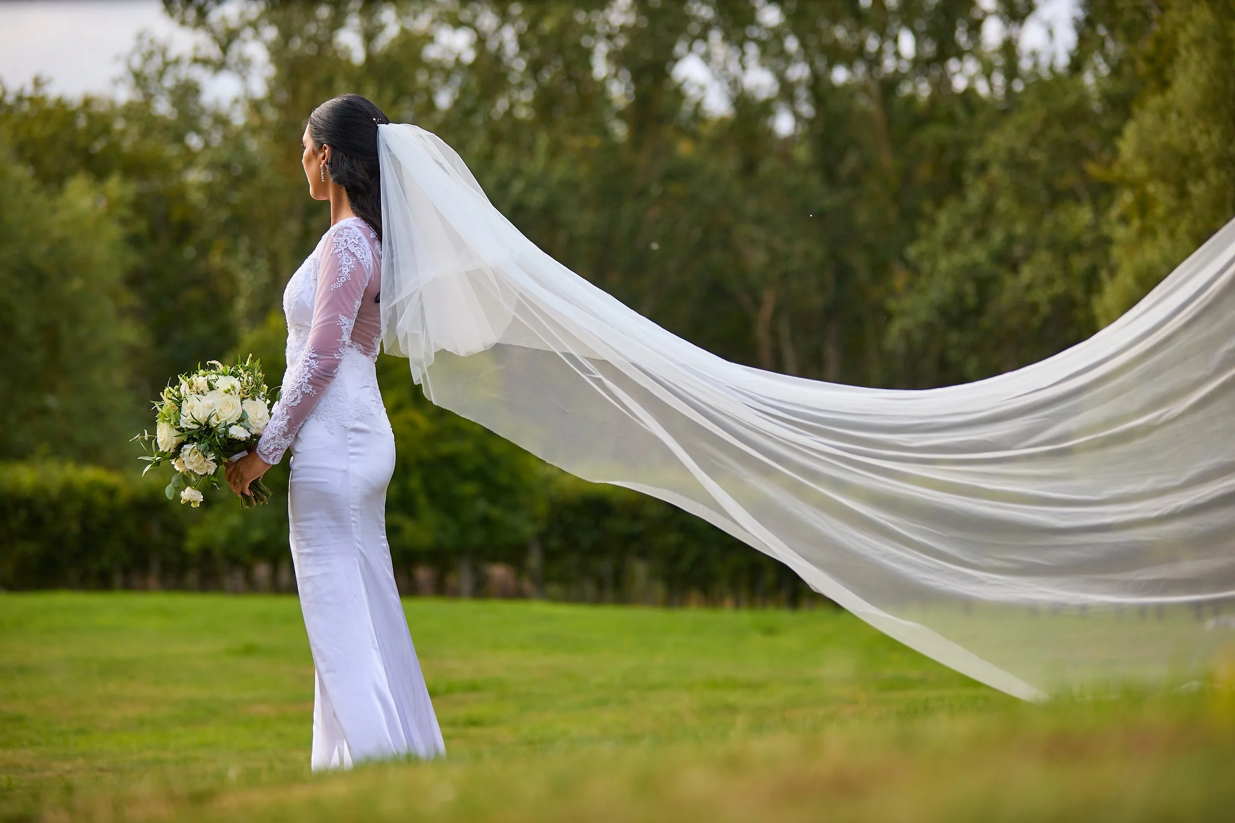A bride in a white wedding dress and veil standing outdoors in a grassy area, holding a bouquet of white roses and greenery. She is facing to the left, with a background of trees and overcast sky.