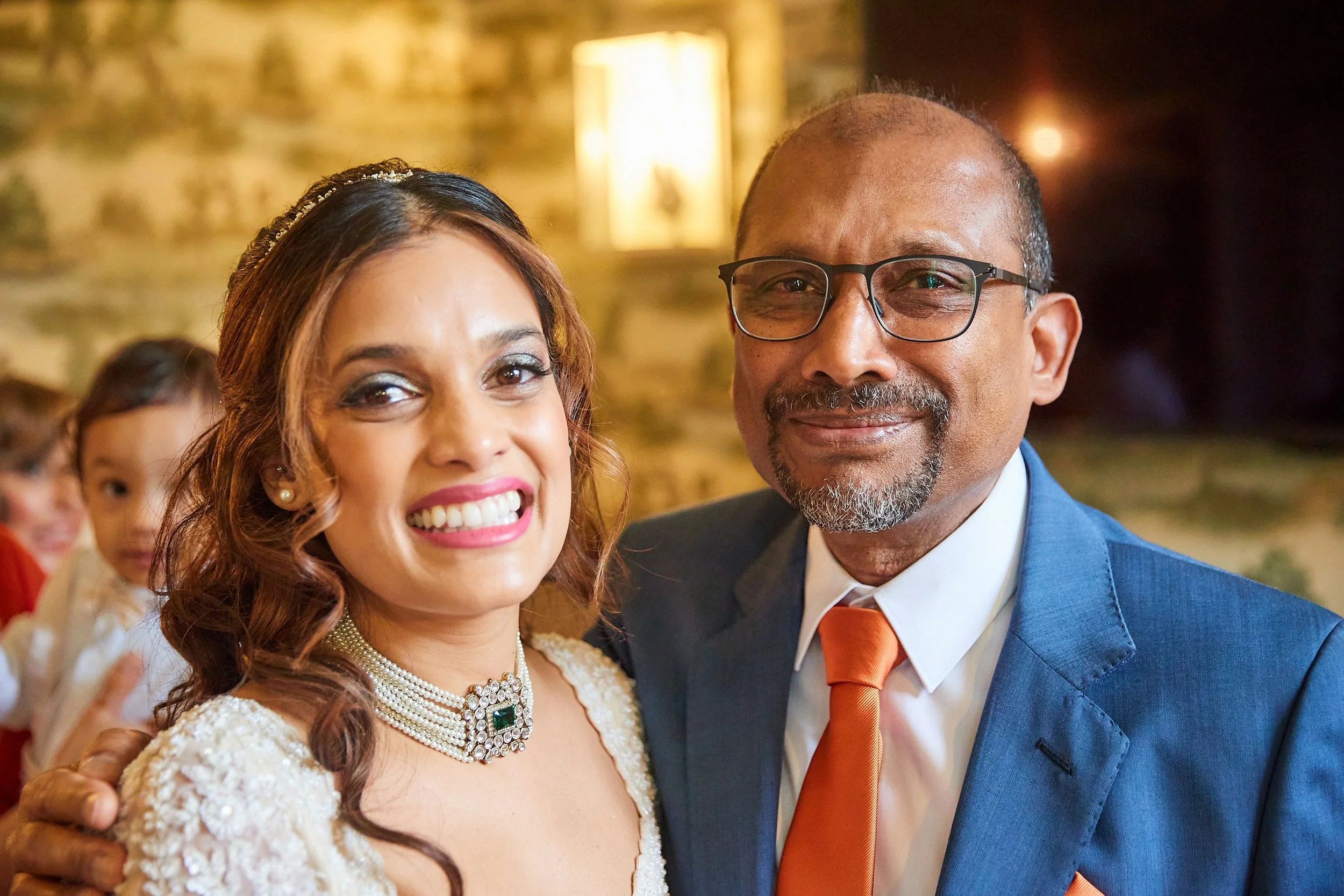 A woman and a man posing together at a social event indoors. The woman is smiling, wearing a white dress and pearl jewelry, and the man is smiling, wearing glasses, a blue suit, and an orange tie. In the background, children are visible, and the sett