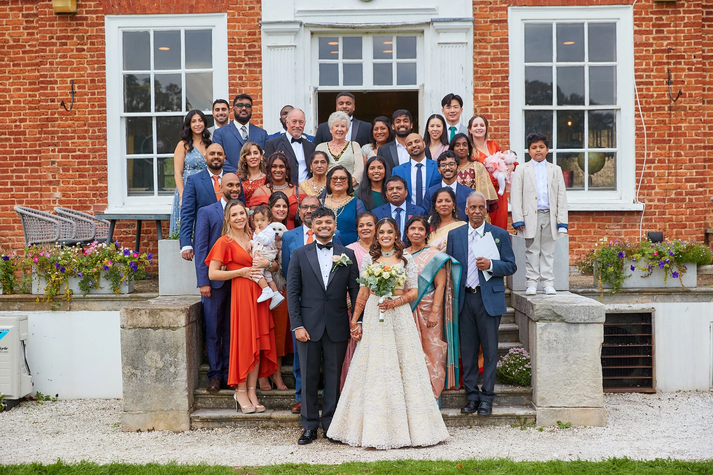 A large group of people dressed in formal attire gathered on the steps outside a brick building with white framing, celebrating a wedding with the bride and groom at the front center. The bride is holding a bouquet of white and green flowers, and the