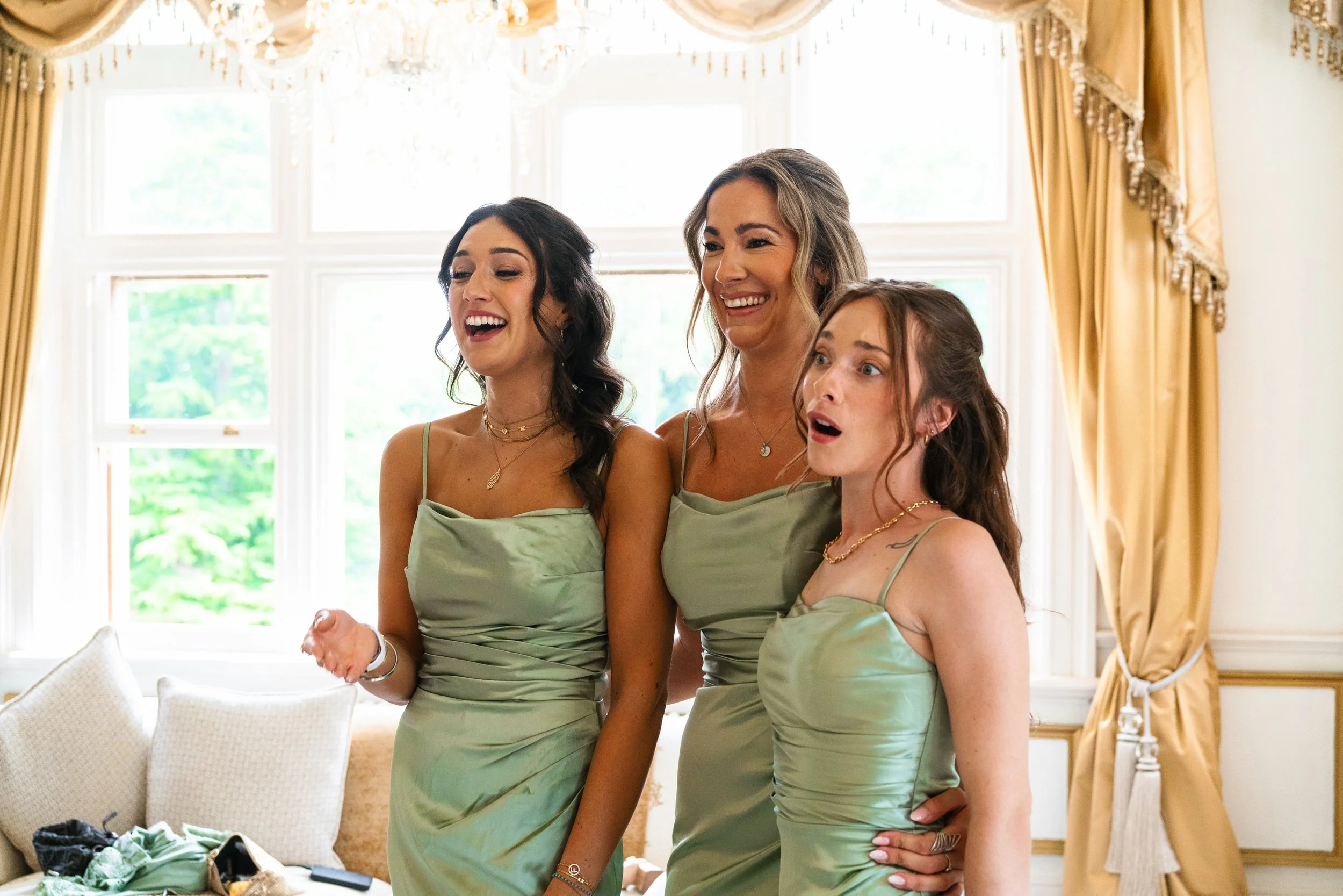 Three women in matching green dresses, smiling and surprised, standing together indoors near a window with gold curtains.