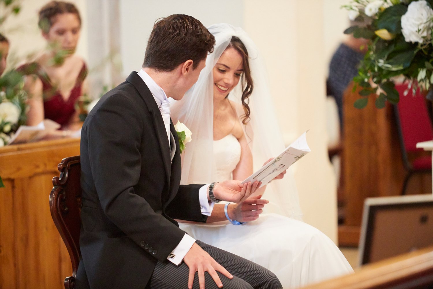 A bride and groom sit during a wedding ceremony, reading vows or a speech, smiling and looking at each other, with guests and floral arrangements in the background.