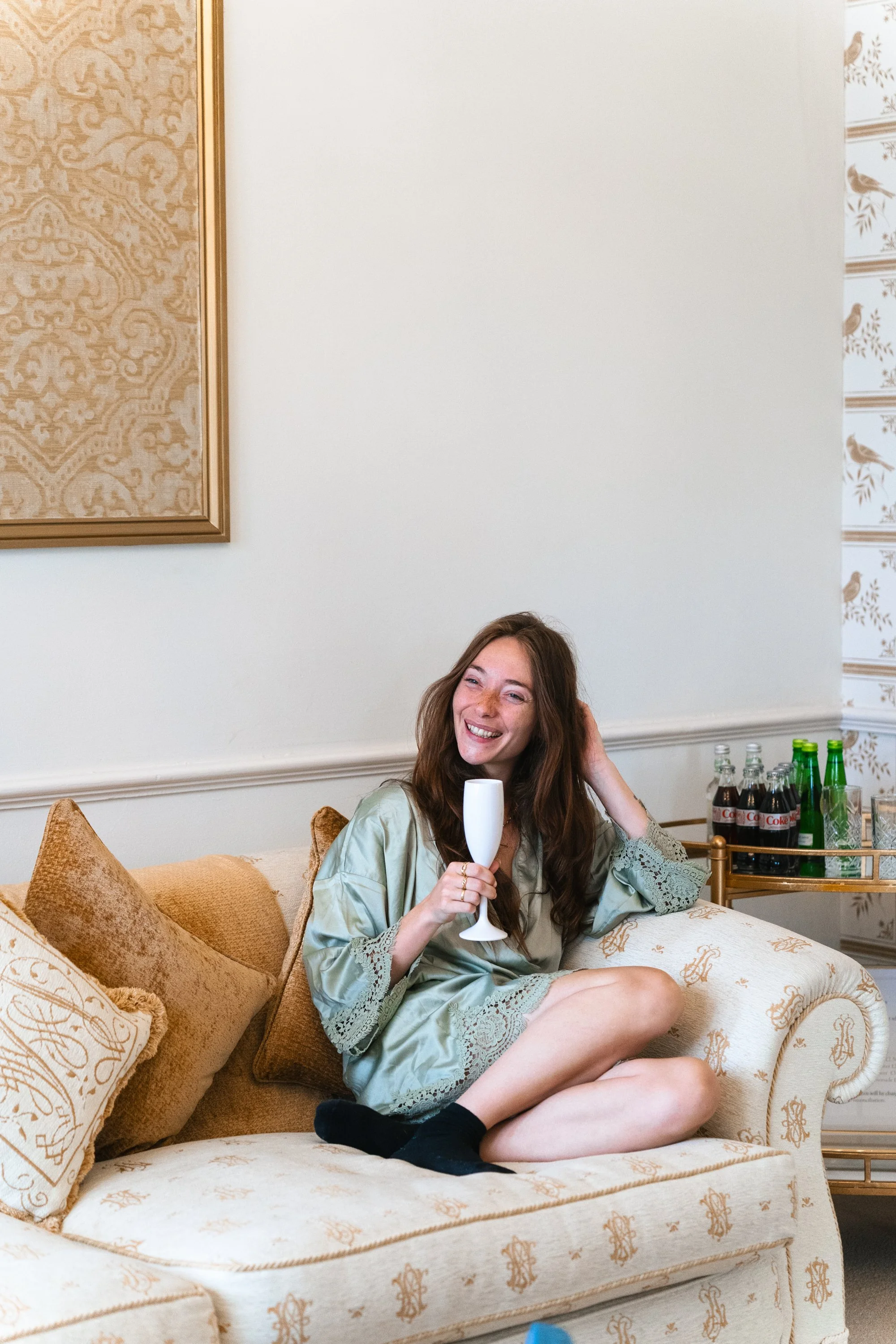 A woman with long brown hair sits on a beige vintage-style sofa holding a white flute glass, smiling, dressed in a silky, light green robe with lace details, surrounded by pillows and drinks on a side cart.