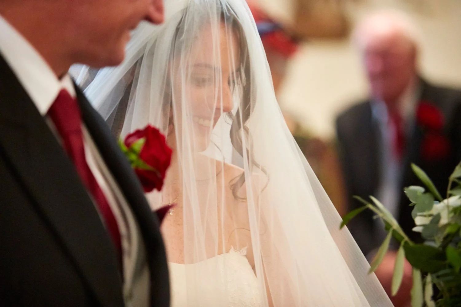 A bride smiling during her wedding ceremony, with a veil covering her face, wearing a white wedding dress, and standing beside her groom in a black suit with a red boutonniere.