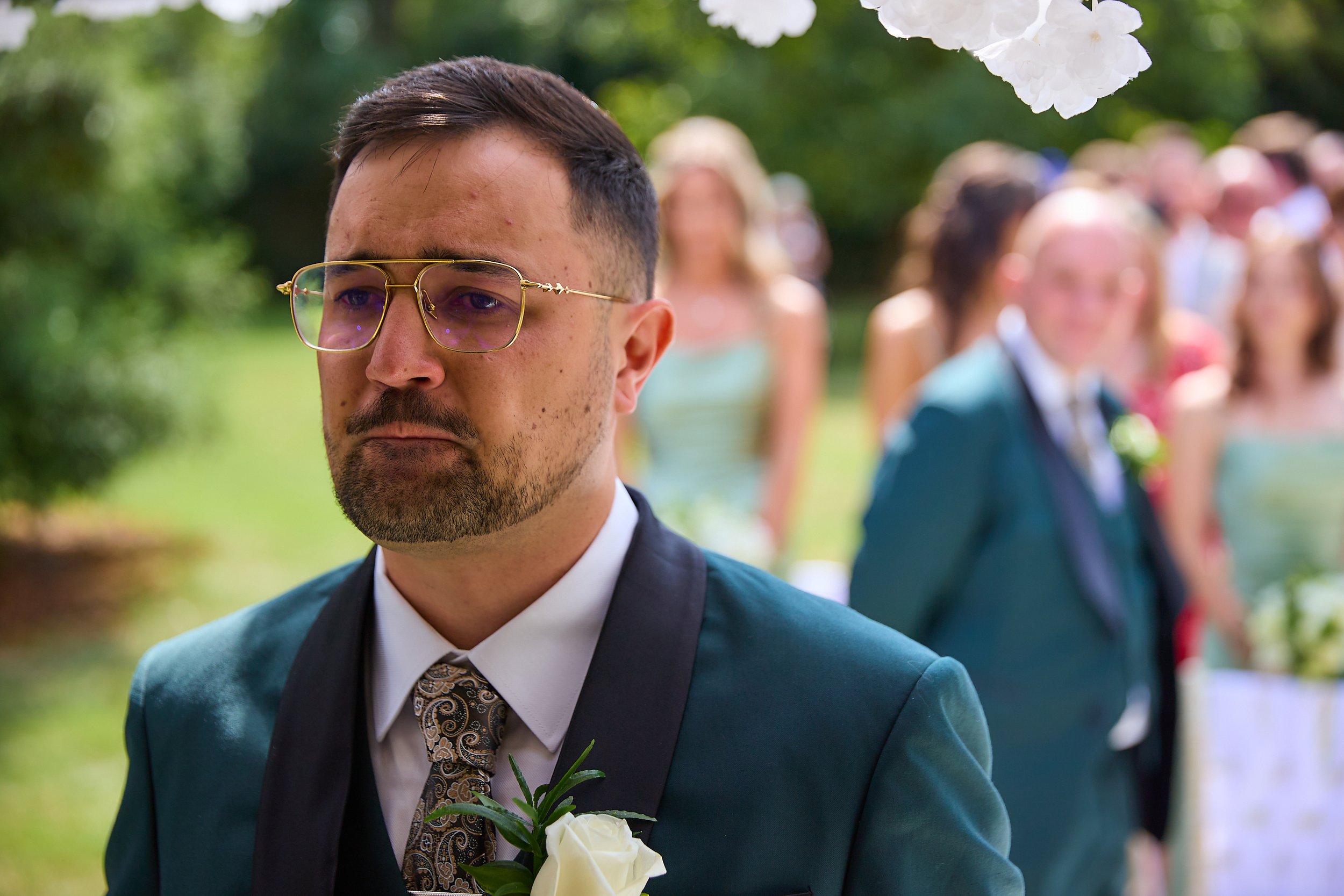 A man in a teal suit with glasses, a patterned tie, and a white shirt, standing outdoors at a wedding ceremony, with a boutonniere of white roses and greenery on his lapel. The background shows other guests, some dressed in formal attire, and greener