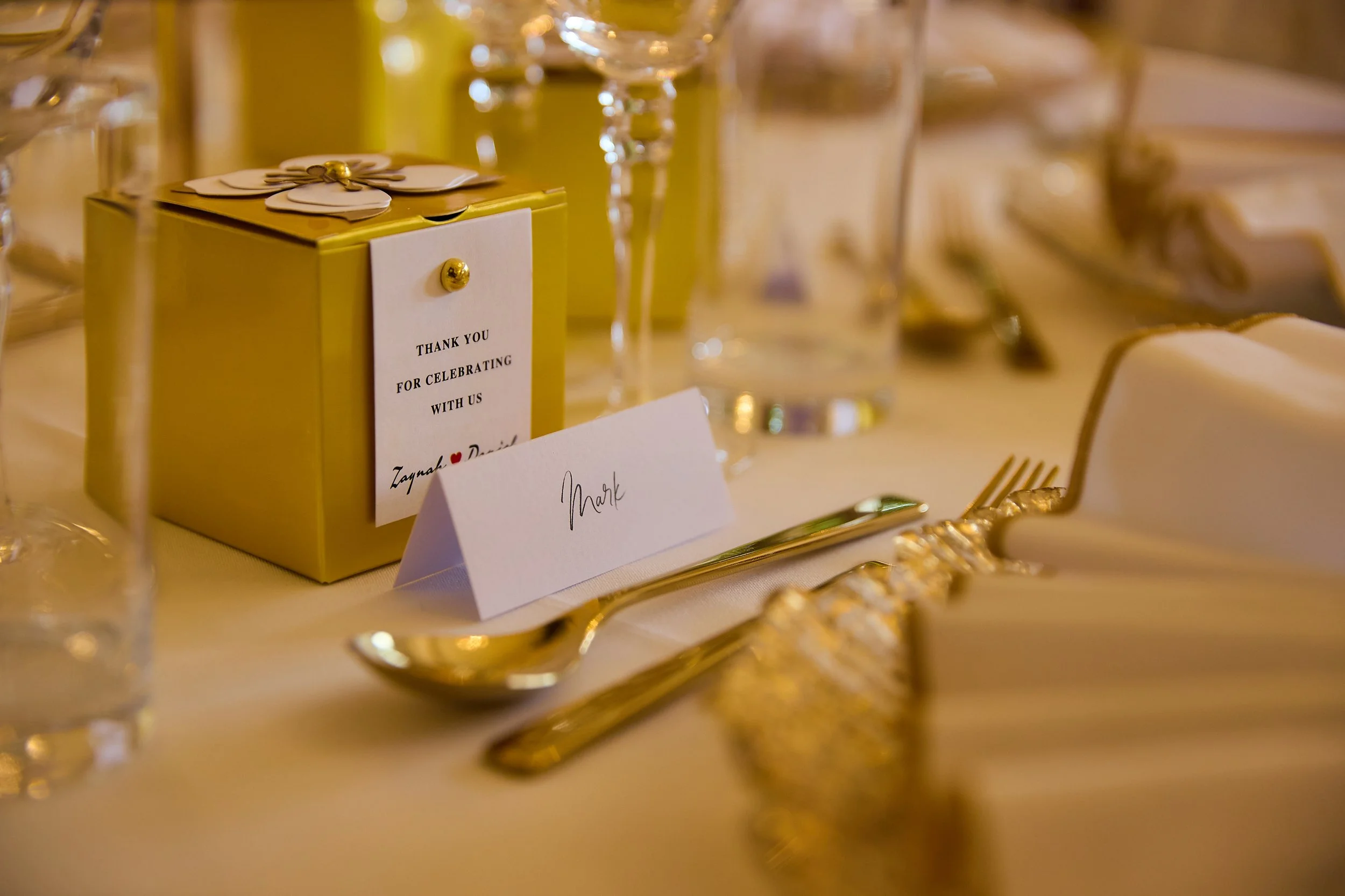 Elegant wedding reception table setting with gold utensils, a white tent card labeled "Mark", and a gold gift box with a thank you note attached.