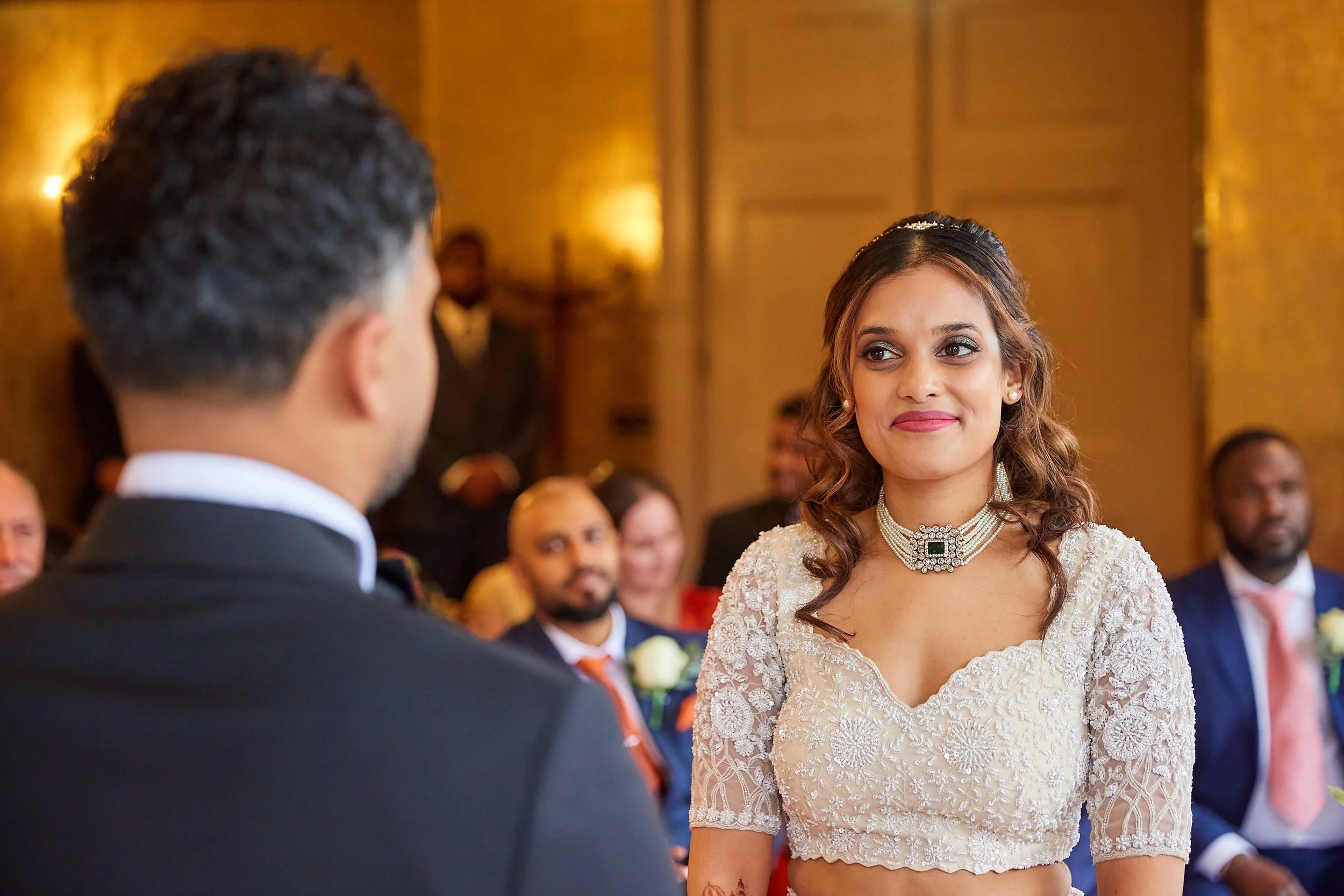 A woman in a wedding dress and jewelry looking at a man in a tuxedo during a wedding ceremony, with guests in the background.