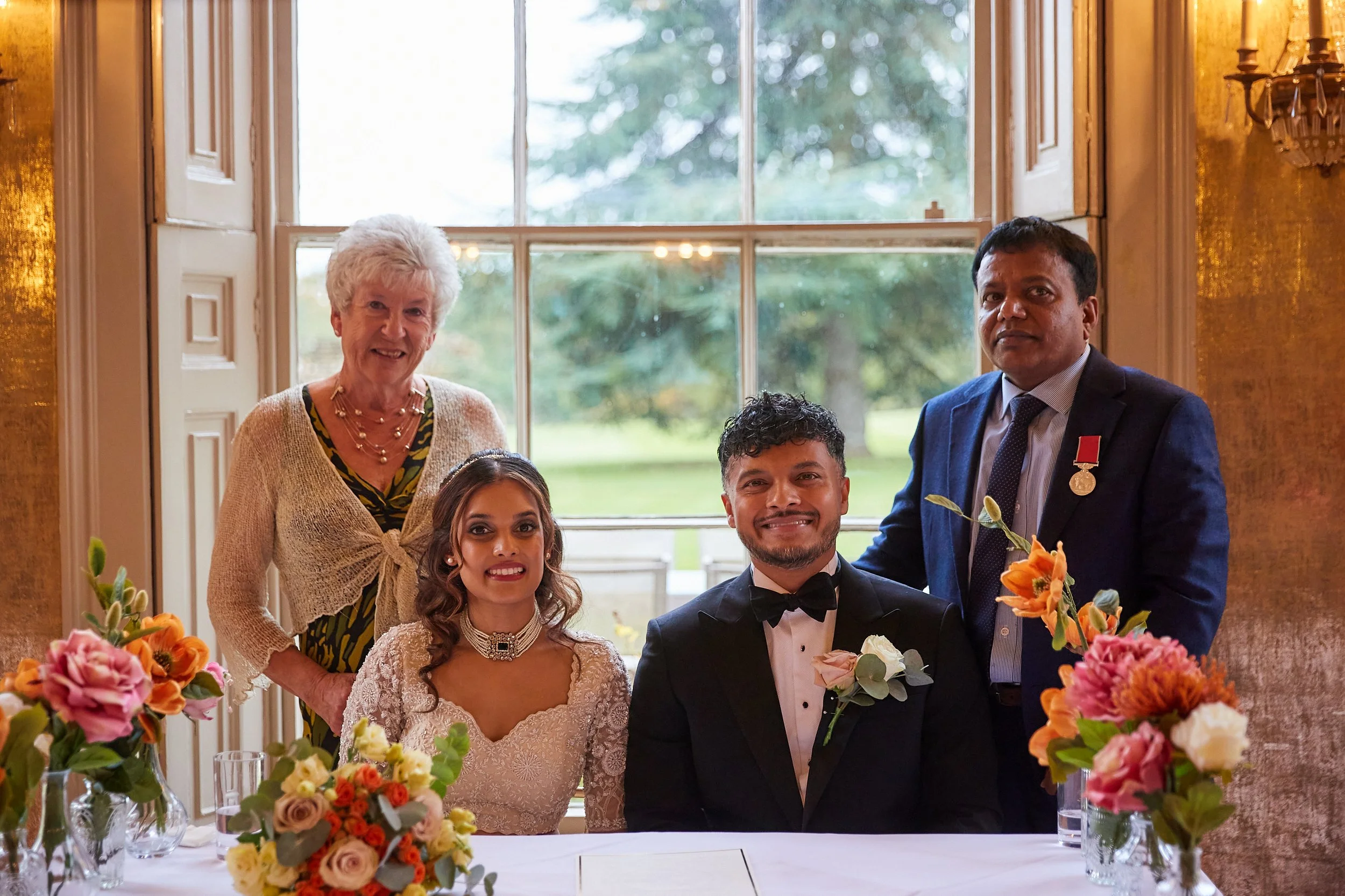 A wedding celebration with five people, including a bride in a white lace dress and a groom in a tuxedo, sitting at a decorated table with floral arrangements, and three standing behind them, inside a brightly lit room with large windows showing gree