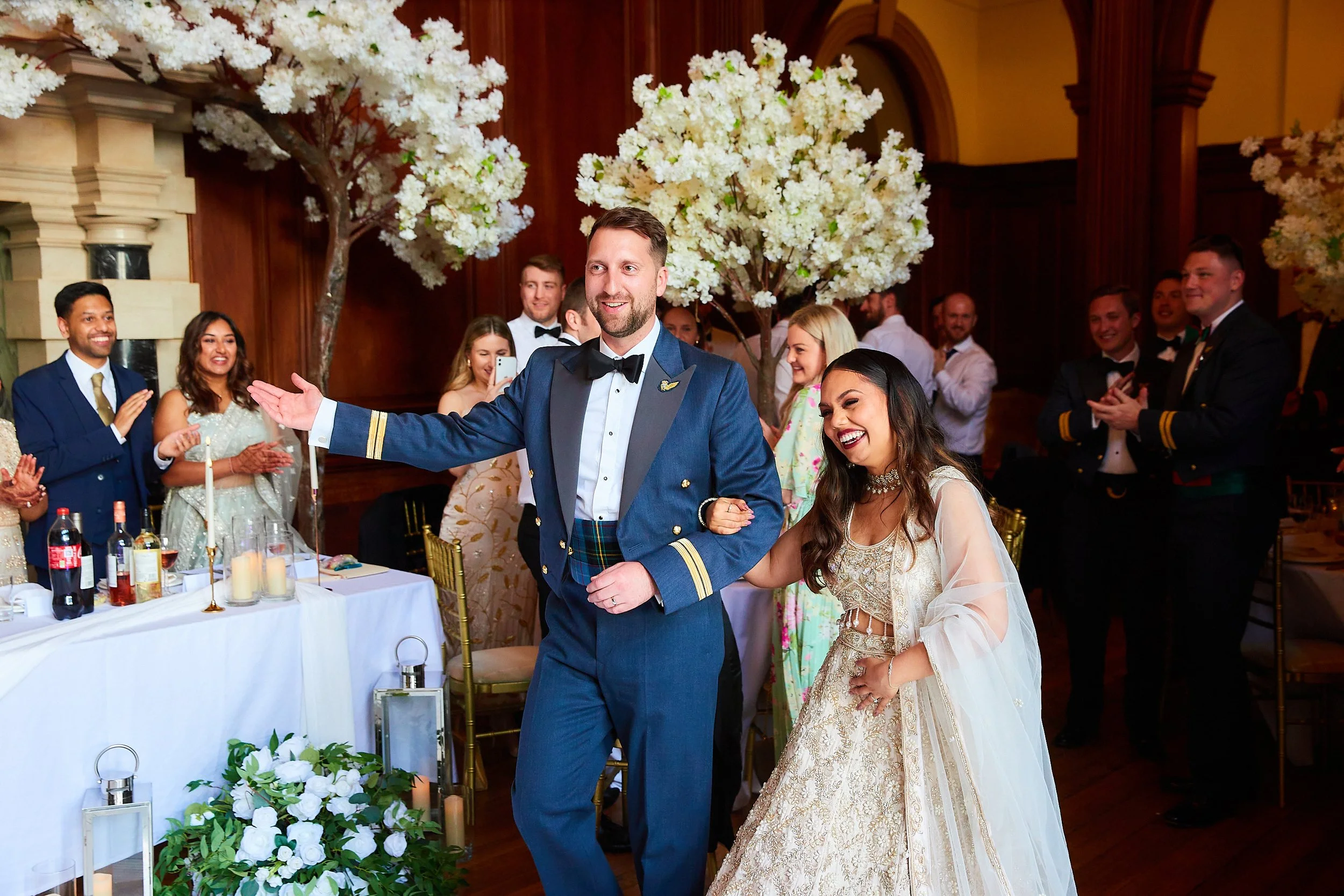 A newlywed couple dancing at their wedding reception, surrounded by guests clapping and smiling indoors with large white floral trees and candles on the tables.